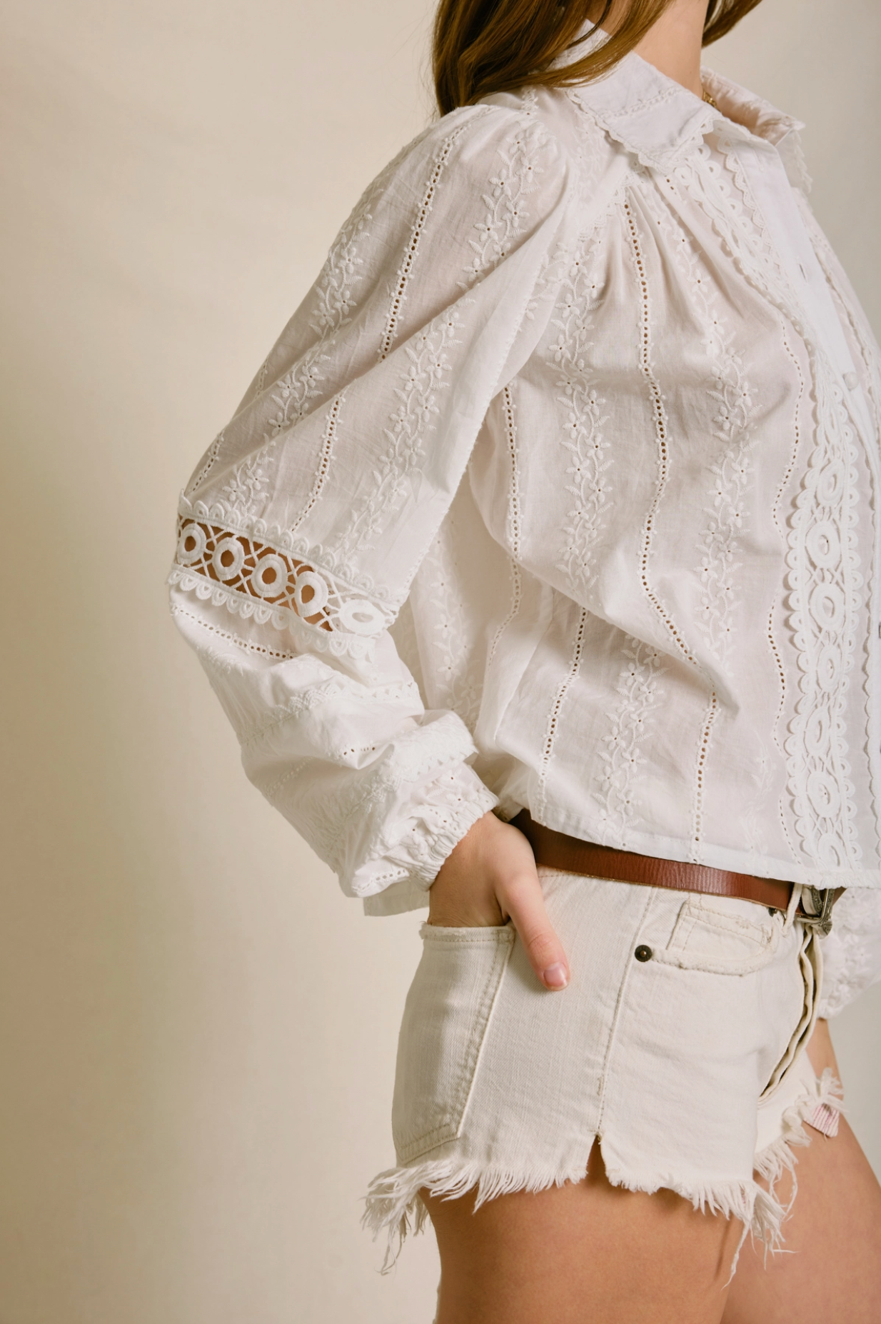 Someone wears the Dress Forum Sunbeam Eyelet Blouse paired with light-colored frayed denim shorts, standing with one hand in a pocket against a neutral background.