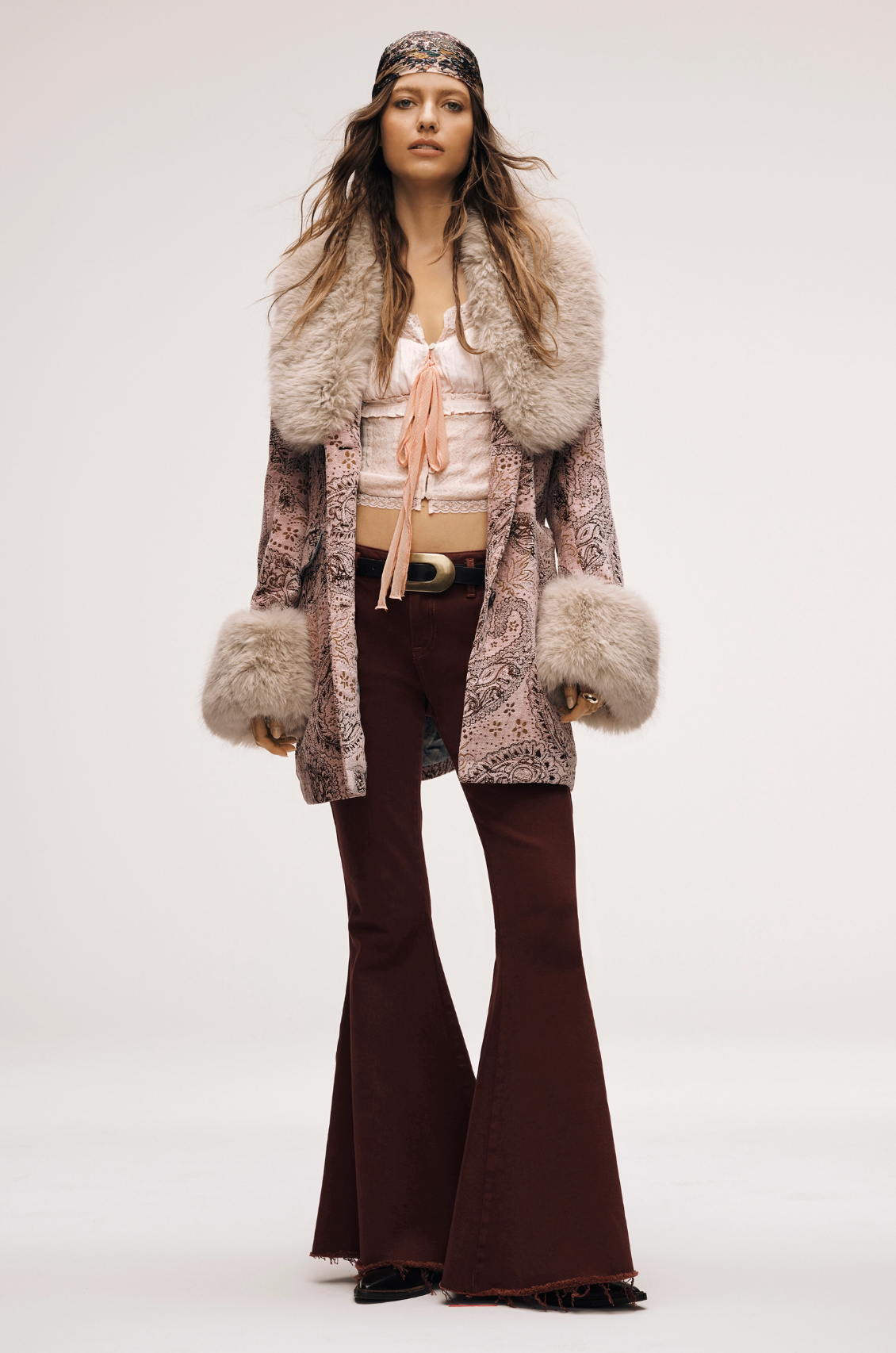A woman models the Free People Mercer Coat, featuring faux fur trim, paired with a white blouse, maroon flared pants, a wide belt, and a patterned headband, capturing a 1970s-inspired bohemian look against a plain background.