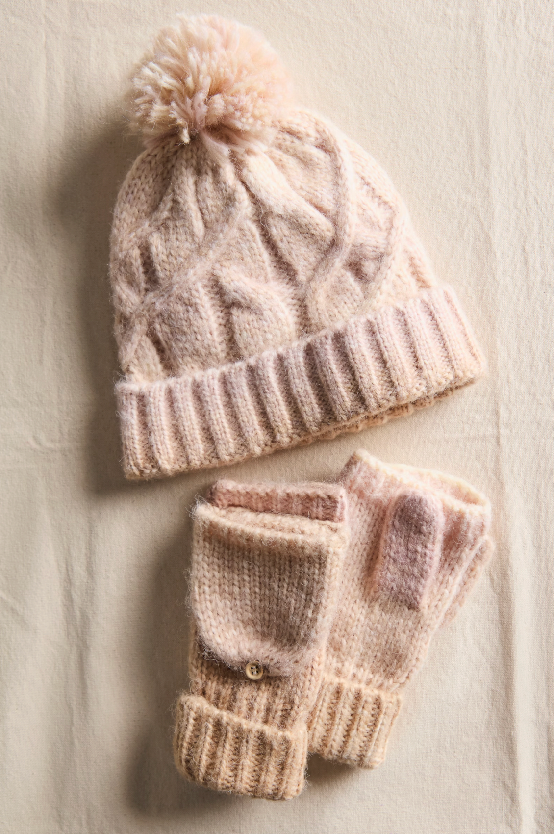 The Rainbow Run Hat and Glove Set in Ivory by Free People, featuring a soft chunky knit beanie with pom-pom and matching fingerless gloves, is shown on a beige fabric background.