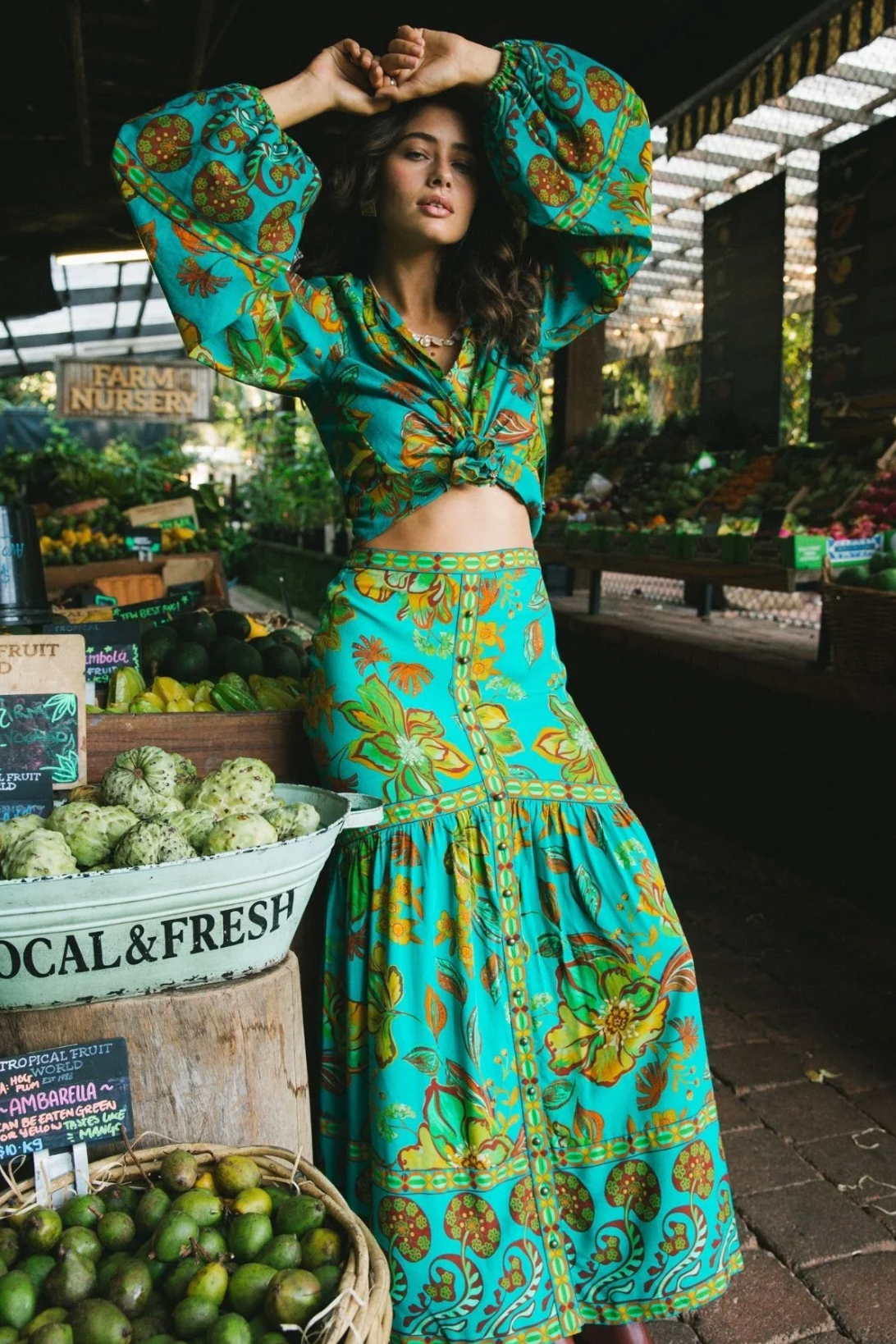 A woman stands in a market wearing the Nine Lives Bazaar Flora Blouse - Peacock with raglan poet sleeves, its vibrant patterns reflecting the colorful tropical fruits as she poses confidently among the fresh produce.