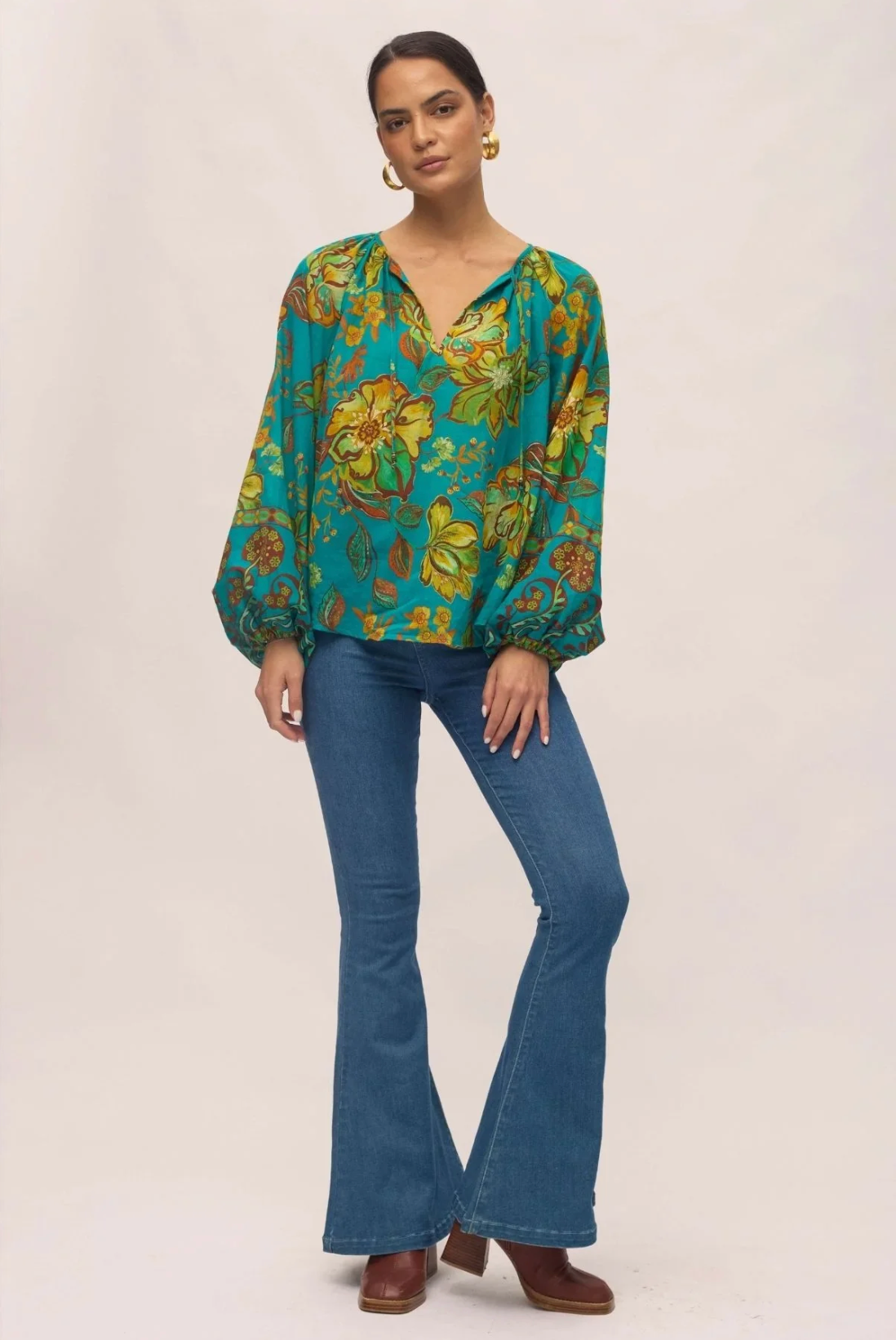 A person poses against a plain light background wearing the Nine Lives Bazaar Flora Blouse in Peacock with raglan poet sleeves, blue flared jeans, brown heeled boots, gold hoop earrings, and their hair pulled back.