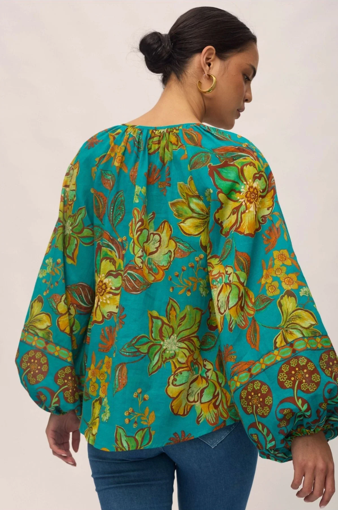 A woman with dark hair in a low bun wears the Nine Lives Bazaar Flora Blouse – Peacock, a teal bohemian top with yellow and green floral patterns and raglan poet sleeves, paired with blue jeans. She is facing away, showing the blouse’s back.