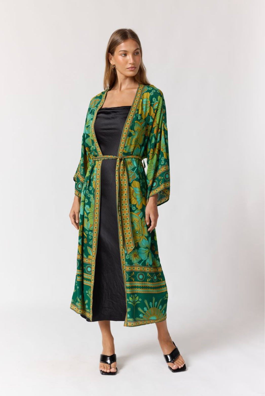 Woman wearing Jade Robe in Emerald with floral patterns over a black dress and black heeled sandals against a white background