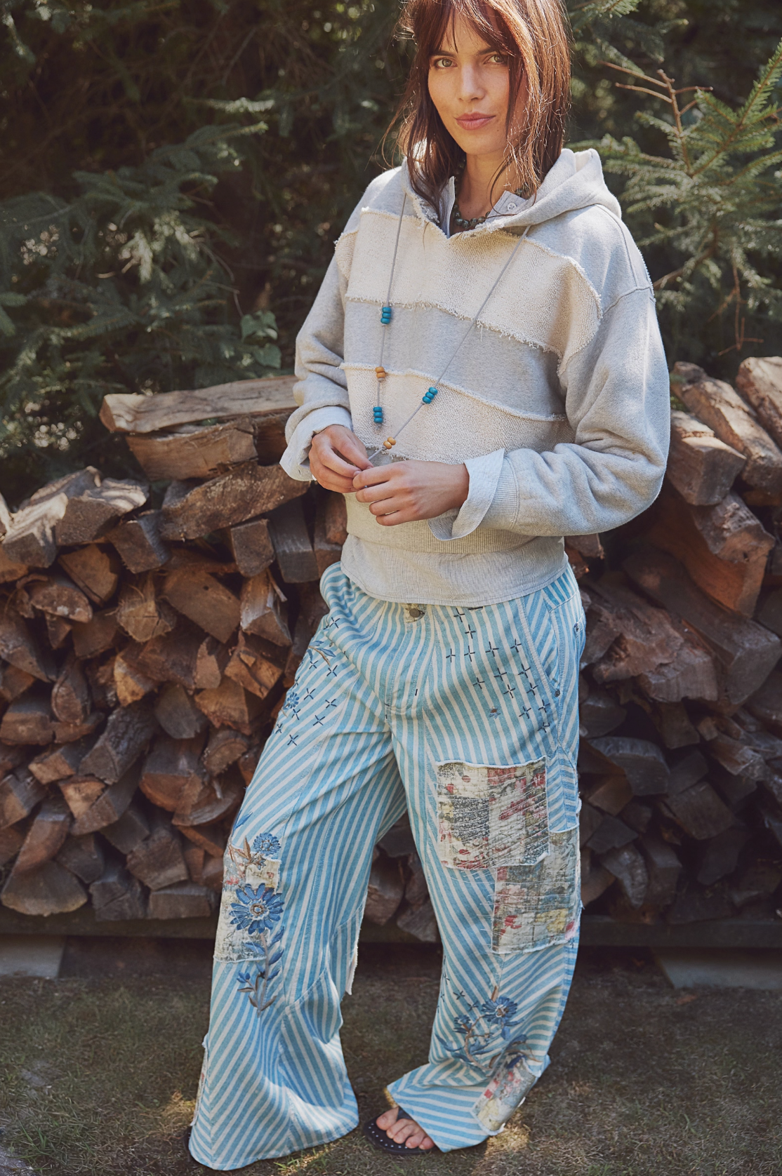 Woman wearing Camp Crush Hoodie with relaxed patchwork pants standing outdoors by stacked firewood