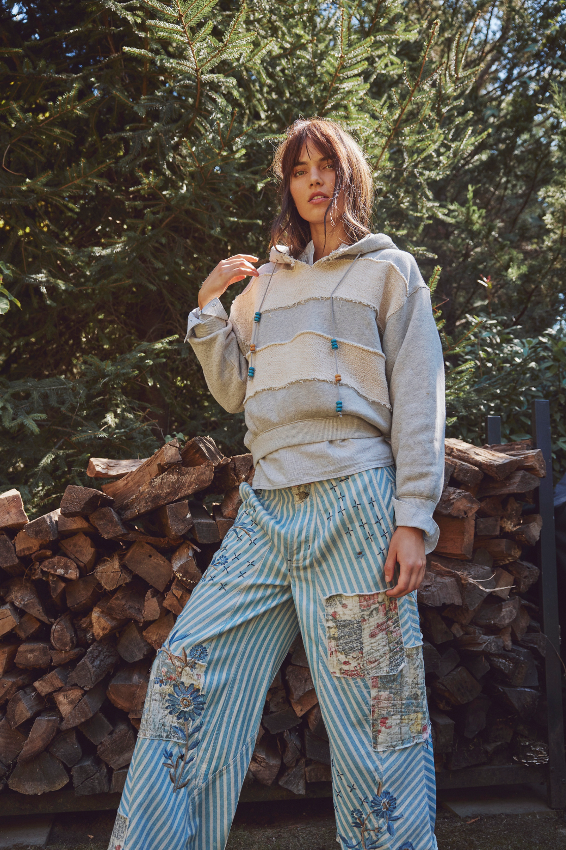Woman wearing a Camp Crush Hoodie outdoors, styled with relaxed patchwork striped pants against stacked firewood.