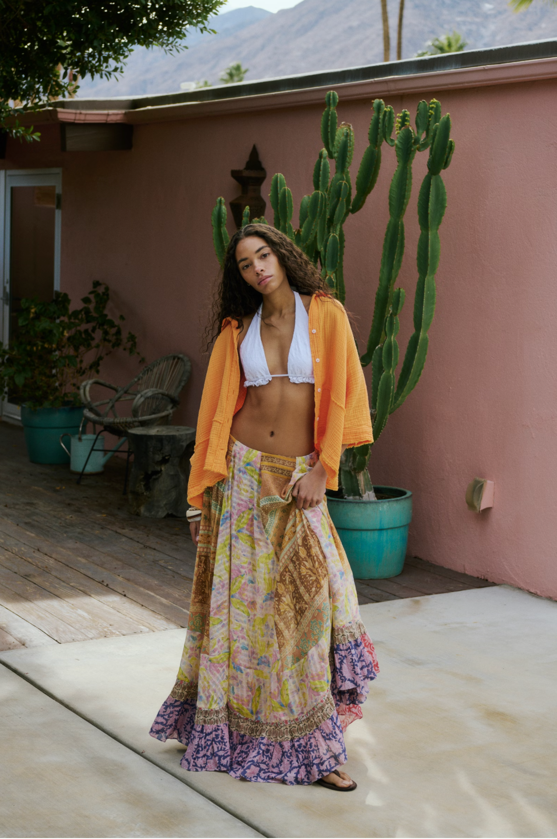 Woman wearing Santa Eulalia Printed Maxi Skirt by Free People with white top and orange shirt outdoors by cactus plant.