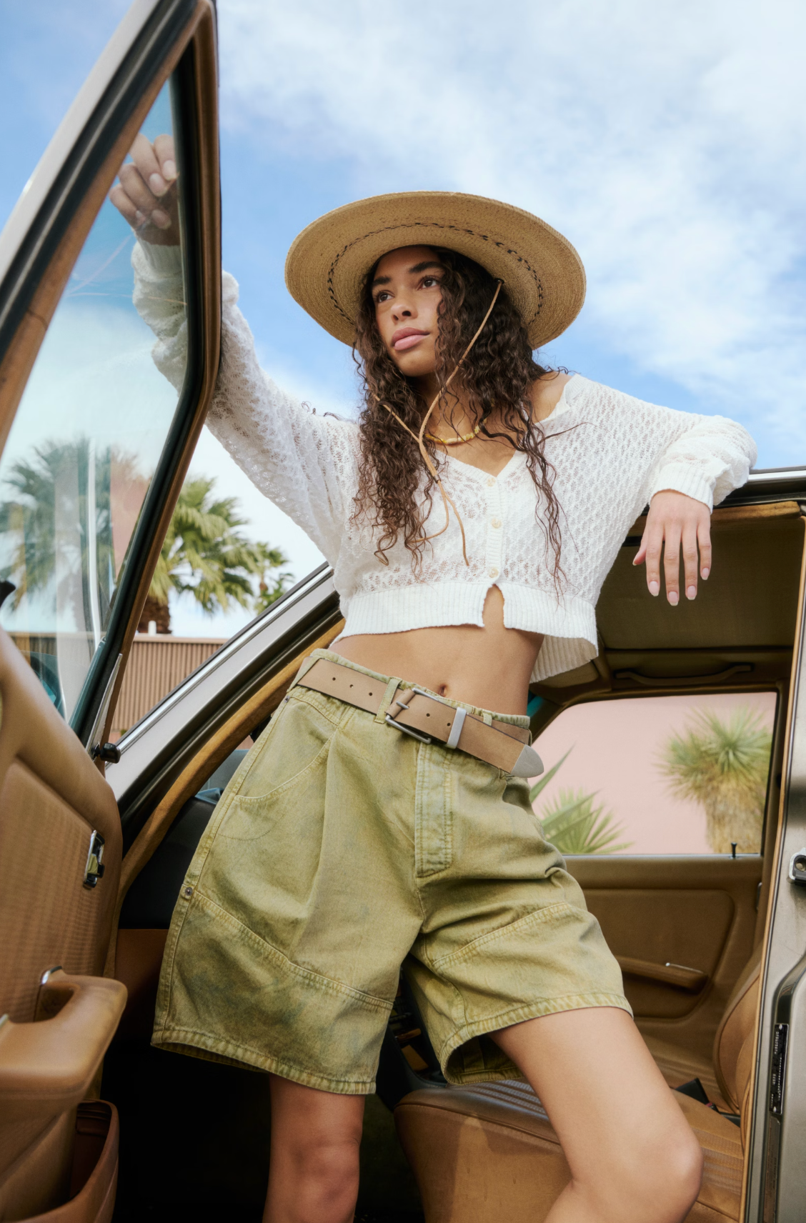 Woman wearing Wild Rebel Short in Outlaw Taupe with a belt and wide-brimmed hat leaning on a car door under blue sky