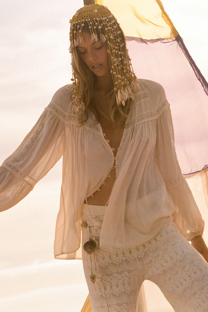 A woman in the Spell Teodora Blouse - White Smoke stands in soft sunlight, lace pants and a decorative headpiece on, as yellow and purple fabric billow translucently behind her.
