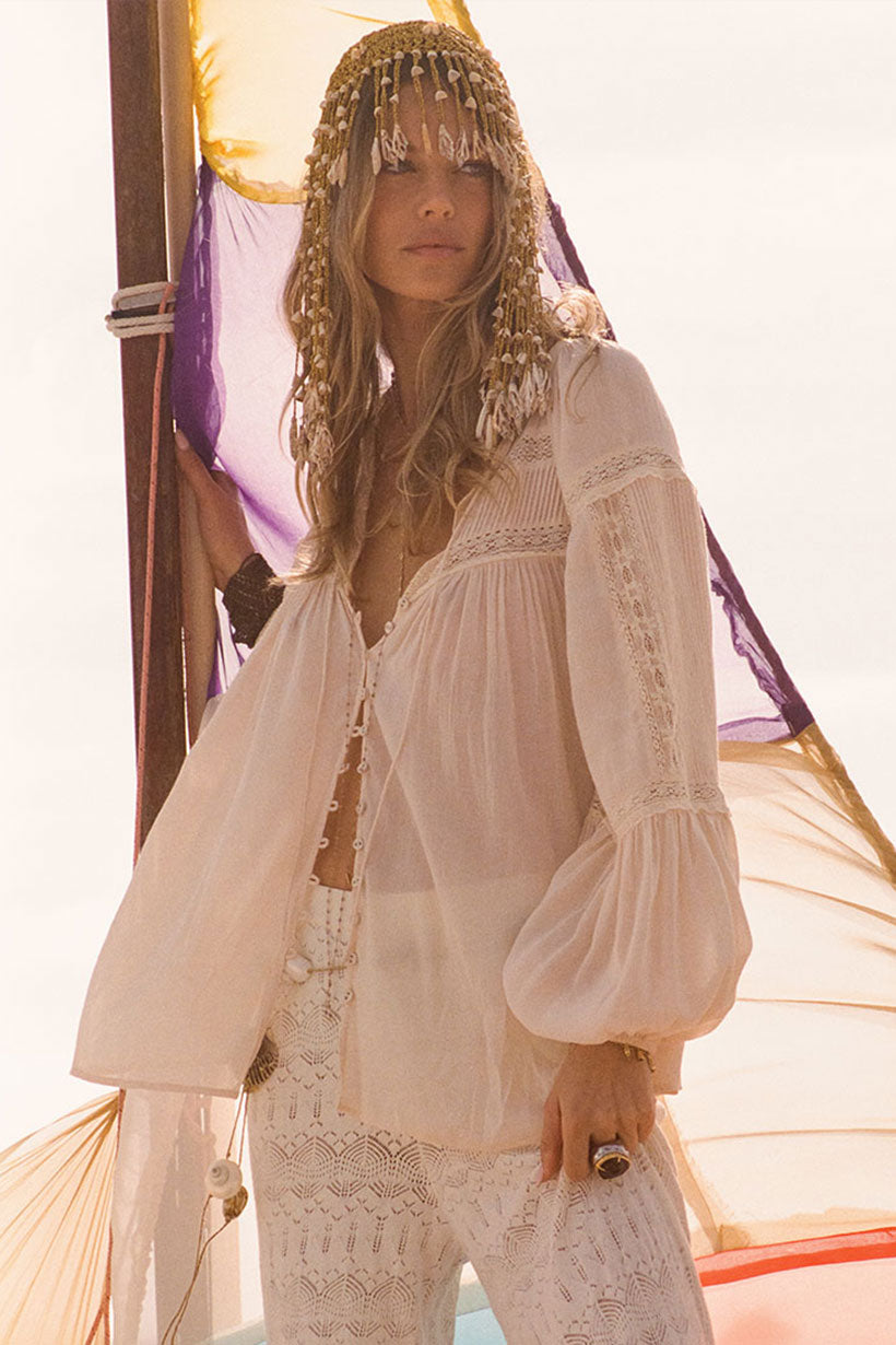 A woman in Spells Teodora Blouse - White Smoke, sheer and long-sleeved, paired with lacy white LENZING ECOVERO Viscose pants and a shell-and-bead headpiece stands outdoors before a sail, softly lit by sunlight.