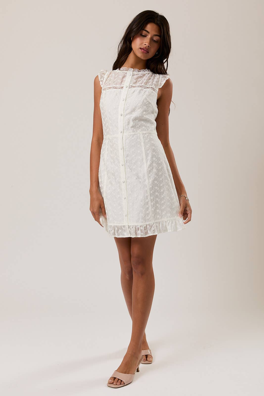 Woman wearing a white Eyelet Lace Mini Dress with delicate buttons, ruffled sleeves, and hem detail in a soft fabric.