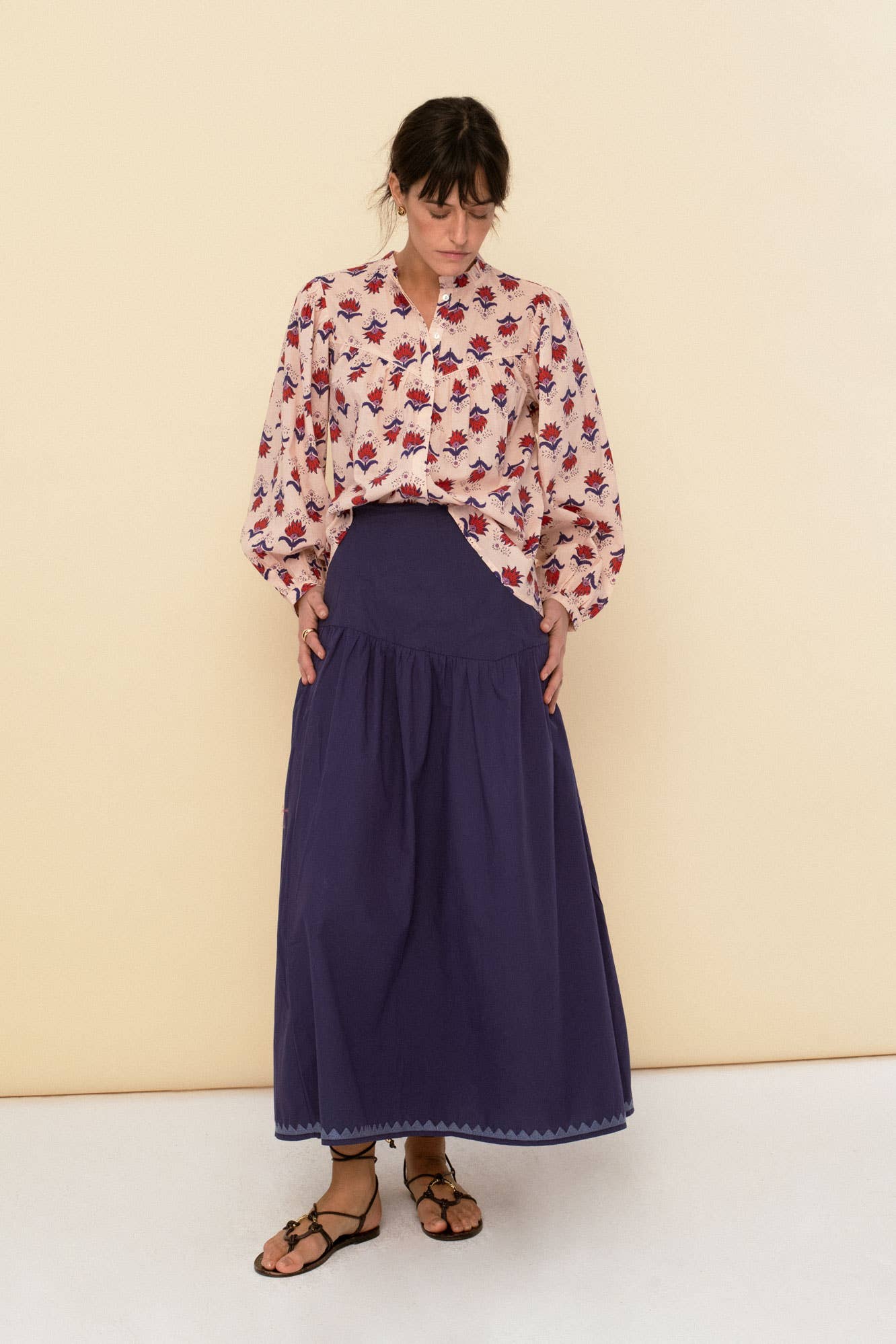 A woman stands against a beige wall, looking down. She wears the Vanessa blouse peach by Sonmer Shop USA and a long, high-waisted navy blue skirt with sandals.