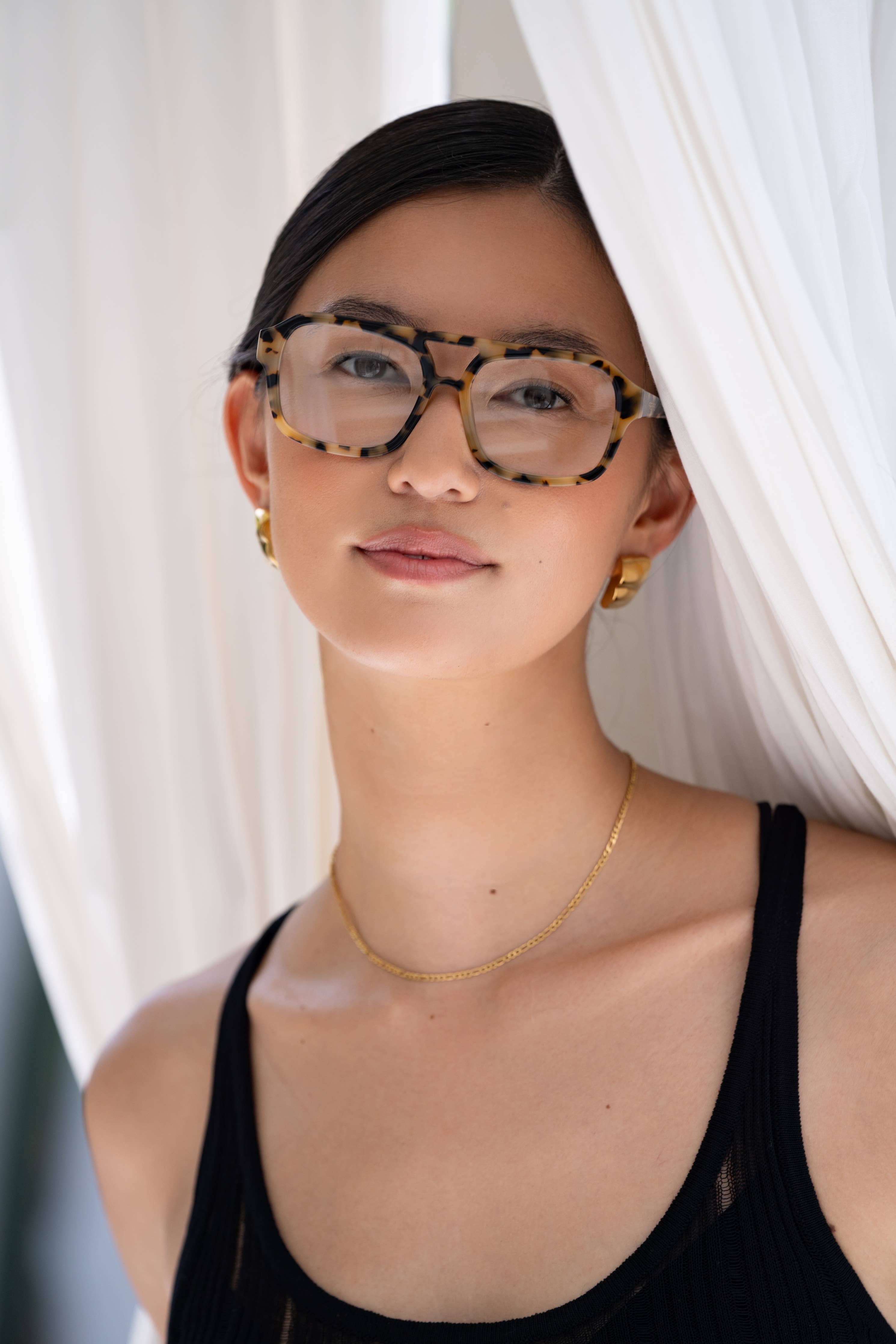 A woman with dark hair, wearing FREYRS Eyewear Havana Blue Light Readers, gold hoop earrings, and a delicate gold necklace stands before white curtains, looking at the camera with a neutral expression.