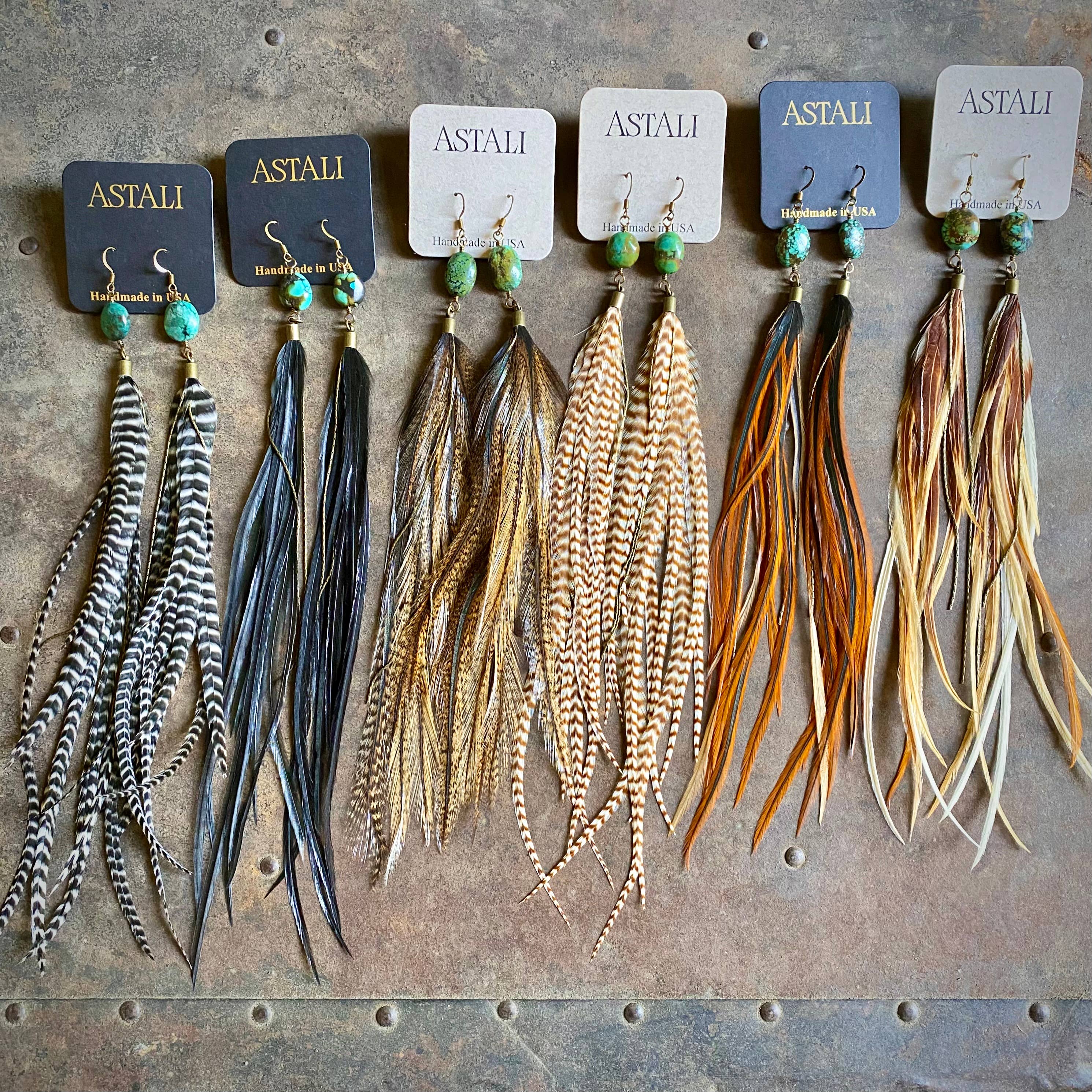 Six pairs of ASTALI Feather & Turquoise Statement Earrings—crafted with antique brass and sustainable coque feathers—are arranged on branded cards atop textured metal, highlighting the unique, eco-friendly jewelry.