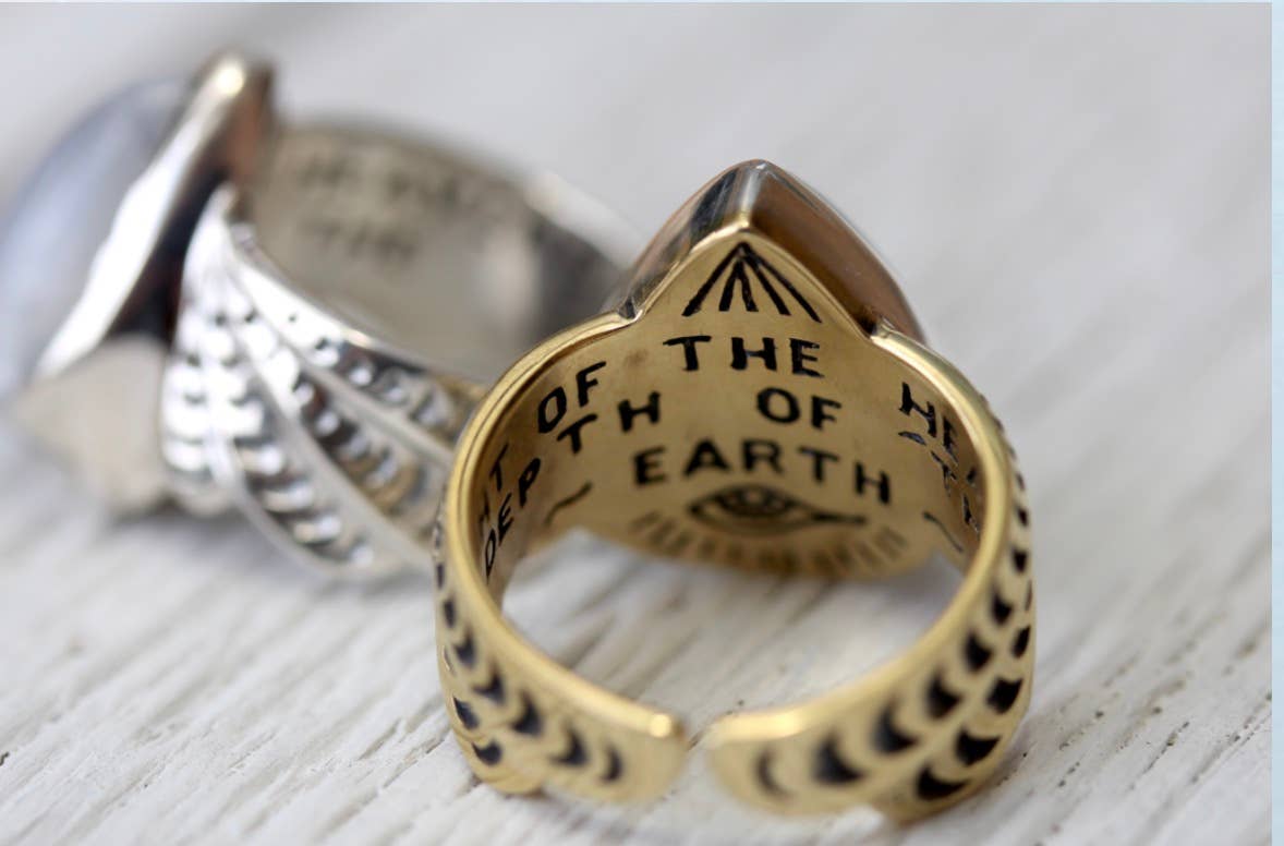 A close-up of two metallic rings on a white surface; the front ring is the Winged Ring by bahgsu jewels, engraved with OF THE H EARTH inside and featuring a feather-like pattern on its band.