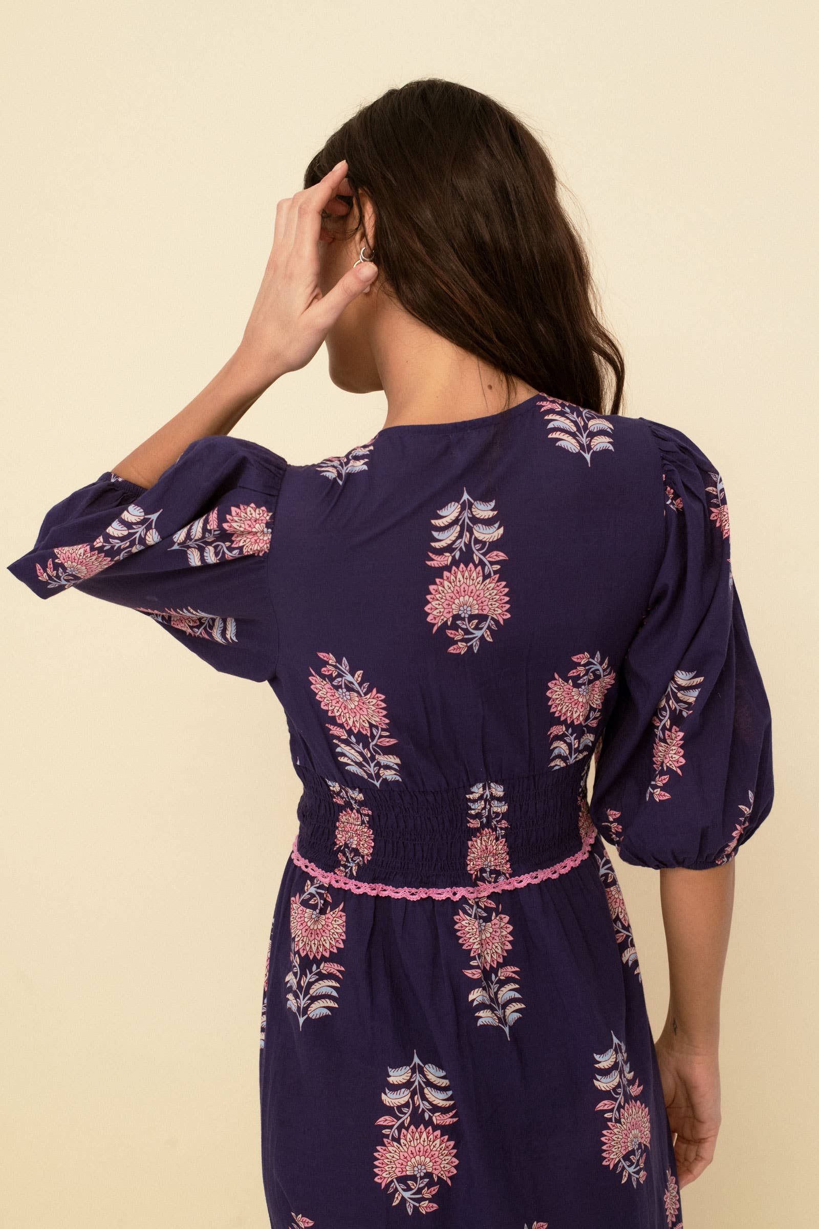 A woman with long brown hair, seen from behind, wears the Magnolia dress blue by Sonmer Shop USA— a romantic vintage navy dress with pink and white florals and puffed sleeves—standing against a plain beige background.