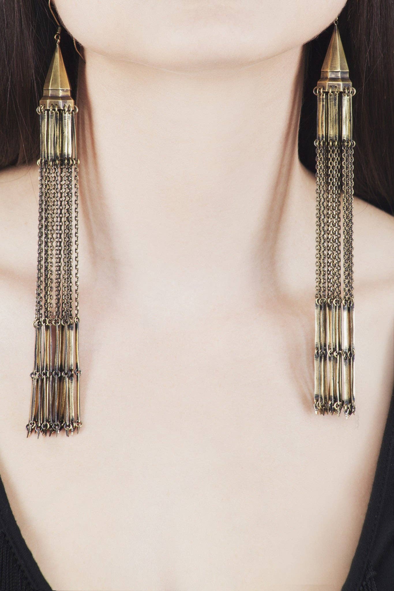 Close-up of a person wearing Jan Hilmer + Sparrow Chandelier earrings—handmade brass earrings with sterling silver hooks and long cylindrical dangles linked by chains, reaching the collarbone, styled with a simple black top.