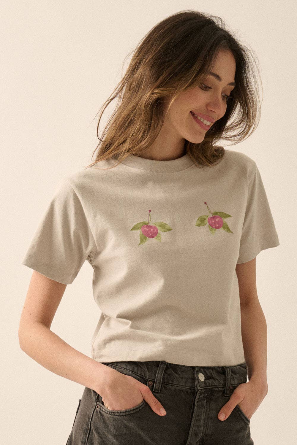 Woman wearing a beige Cherries Cropped Garment-Dyed Graphic Tee with cherry graphics and black jeans, smiling confidently.