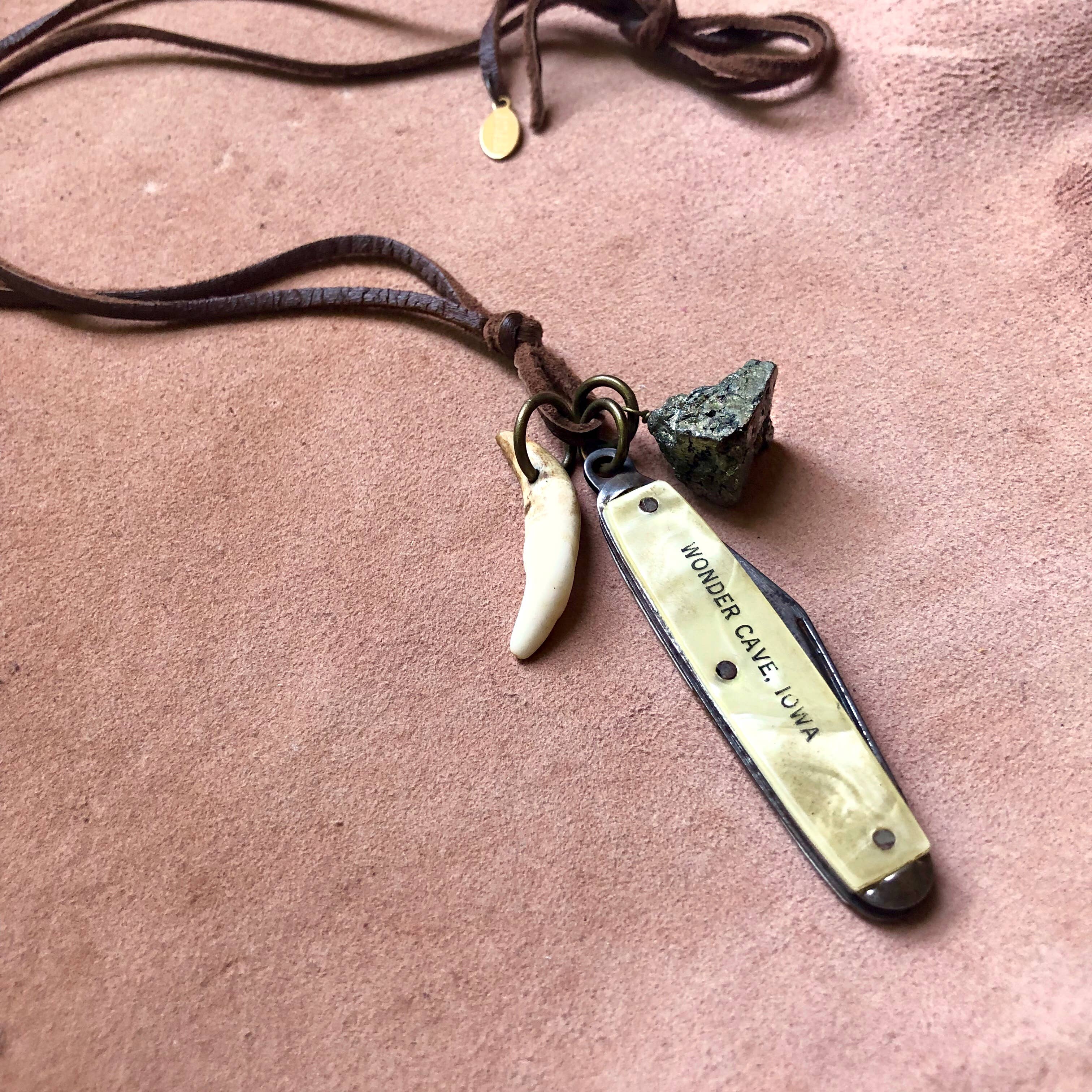 ASTALI’s Vintage Pocket Knife & Coyote Tooth Necklace features a pyrite pendant and deerskin leather, with a coyote tooth and engraved pocket knife charms displayed on a soft pink surface.