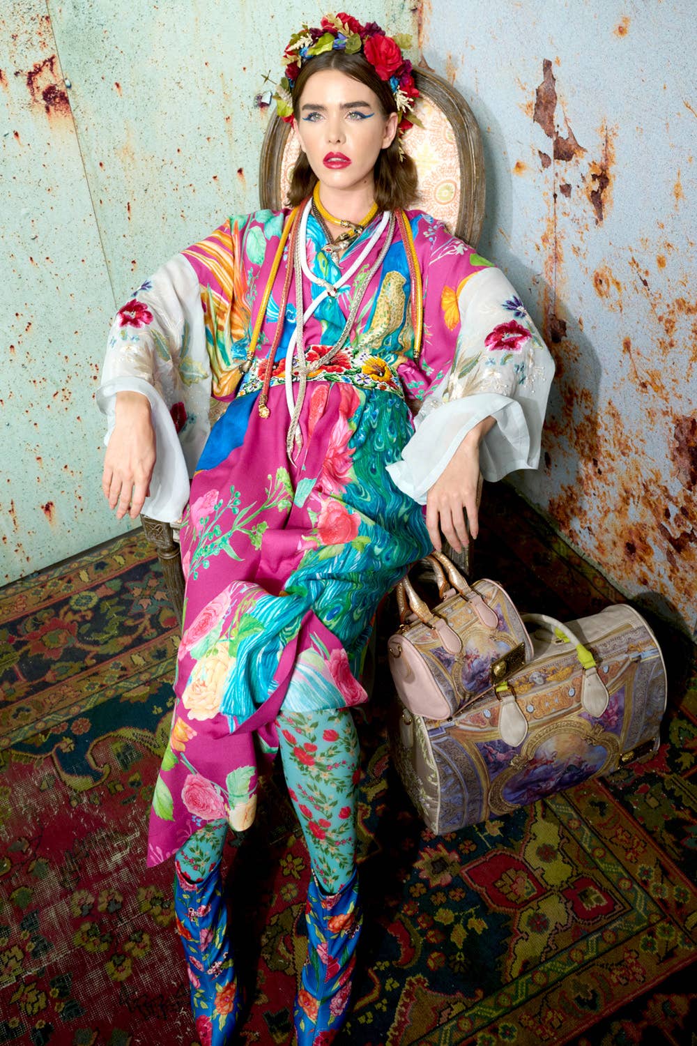A woman in the Marie Antoinette Kimono by Aratta, featuring embroidered floral sleeves, sits on a chair against a rustic wall. She wears a flower crown and bright makeup, with a patterned bag and rug adding to the bold, eclectic look.