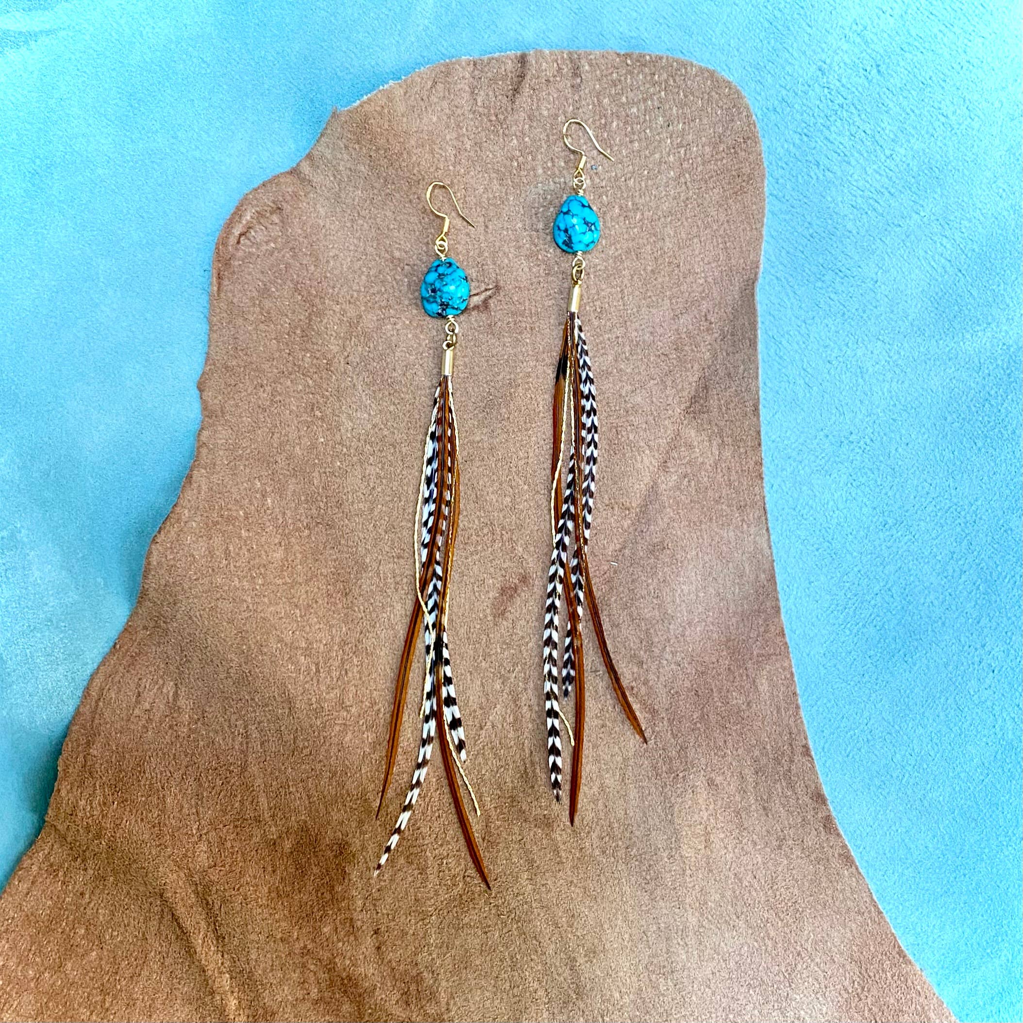 ASTALI’s Turquoise & Mini Feather Earrings – Grizzly & Fawn/Gold feature gold-plated turquoise with brown and black patterned feathers, shown on a soft tan and blue textured background.