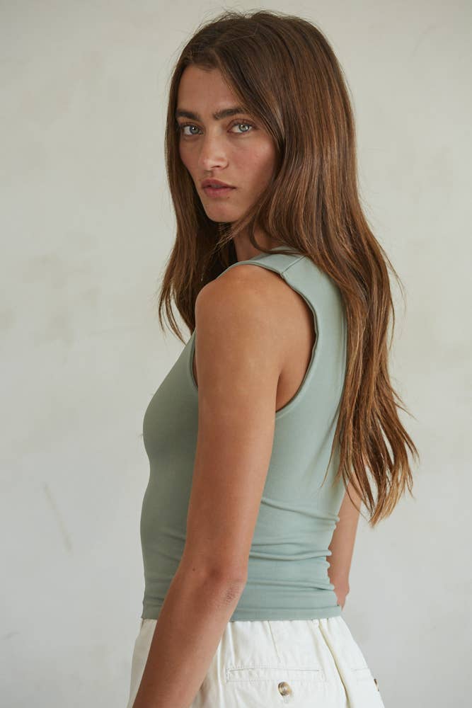 A woman with long brown hair wears a light green By Together Double Layered Seamless Top and white pants, standing sideways and looking at the camera against a plain, light-colored background.