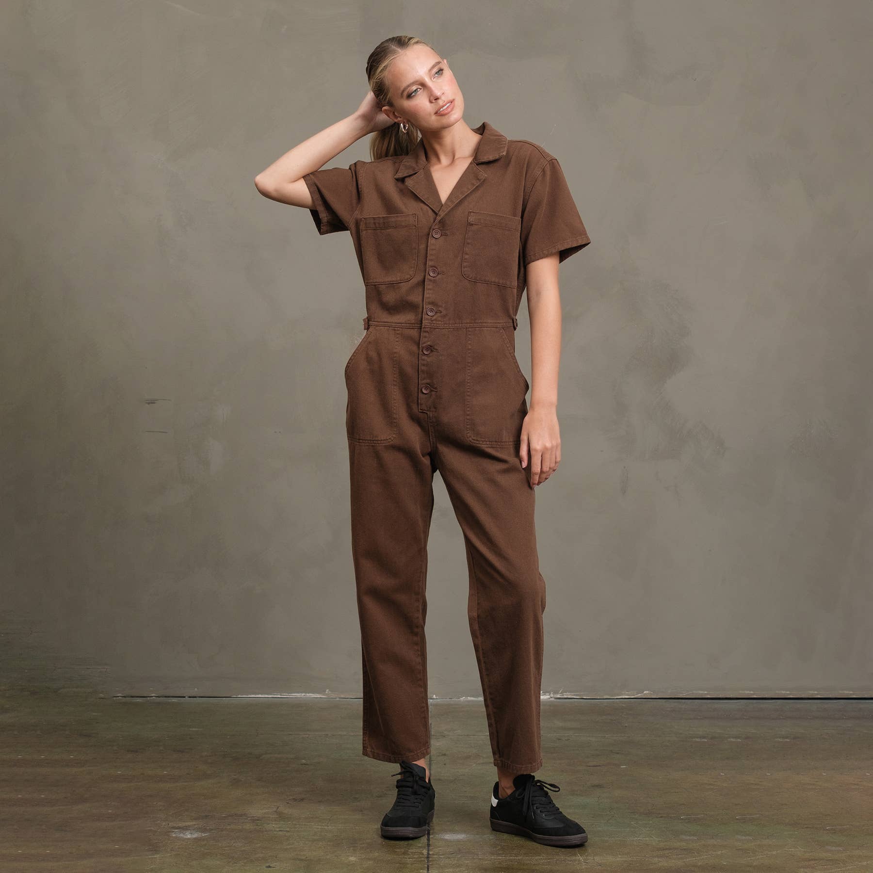 A person wearing the RAISONNEL Field Jumpsuit (RJP1001) and black sneakers stands against a gray wall, one hand touching the back of their head and their head tilted to the side.