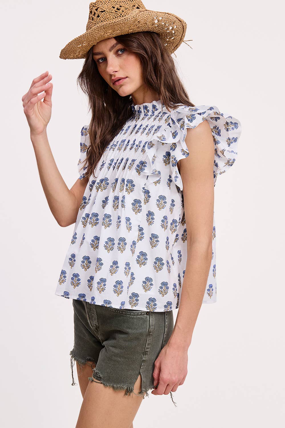 A woman wears the Ultramarine Celeste Flutter Sleeve Blouse with blue floral prints and olive green denim shorts, paired with a straw hat, standing against a plain light background.