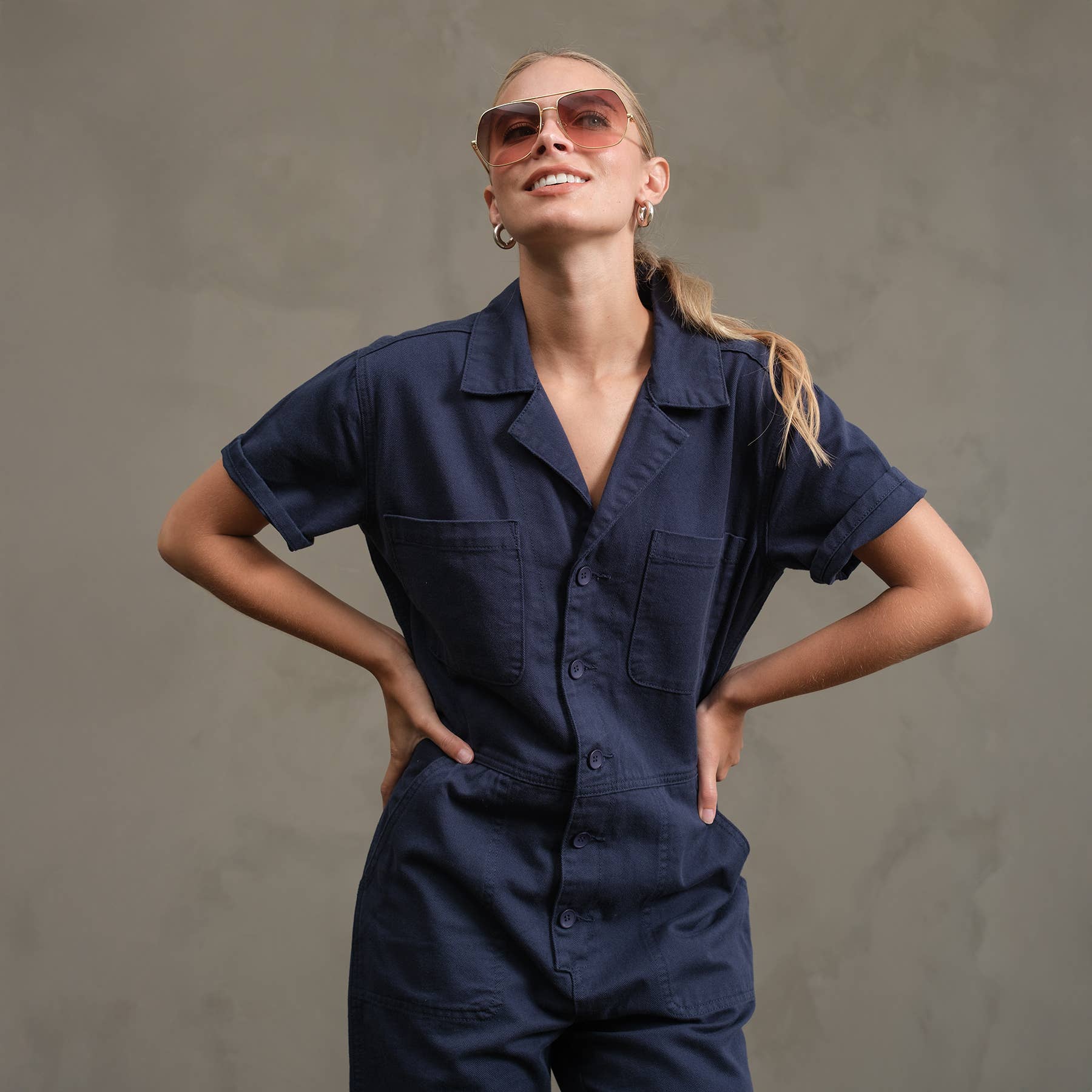 A person wears the RAISONNEL Field Jumpsuit - RJP1001 in navy blue with sunglasses, standing against a plain gray background, hands on hips and smiling while looking upward.