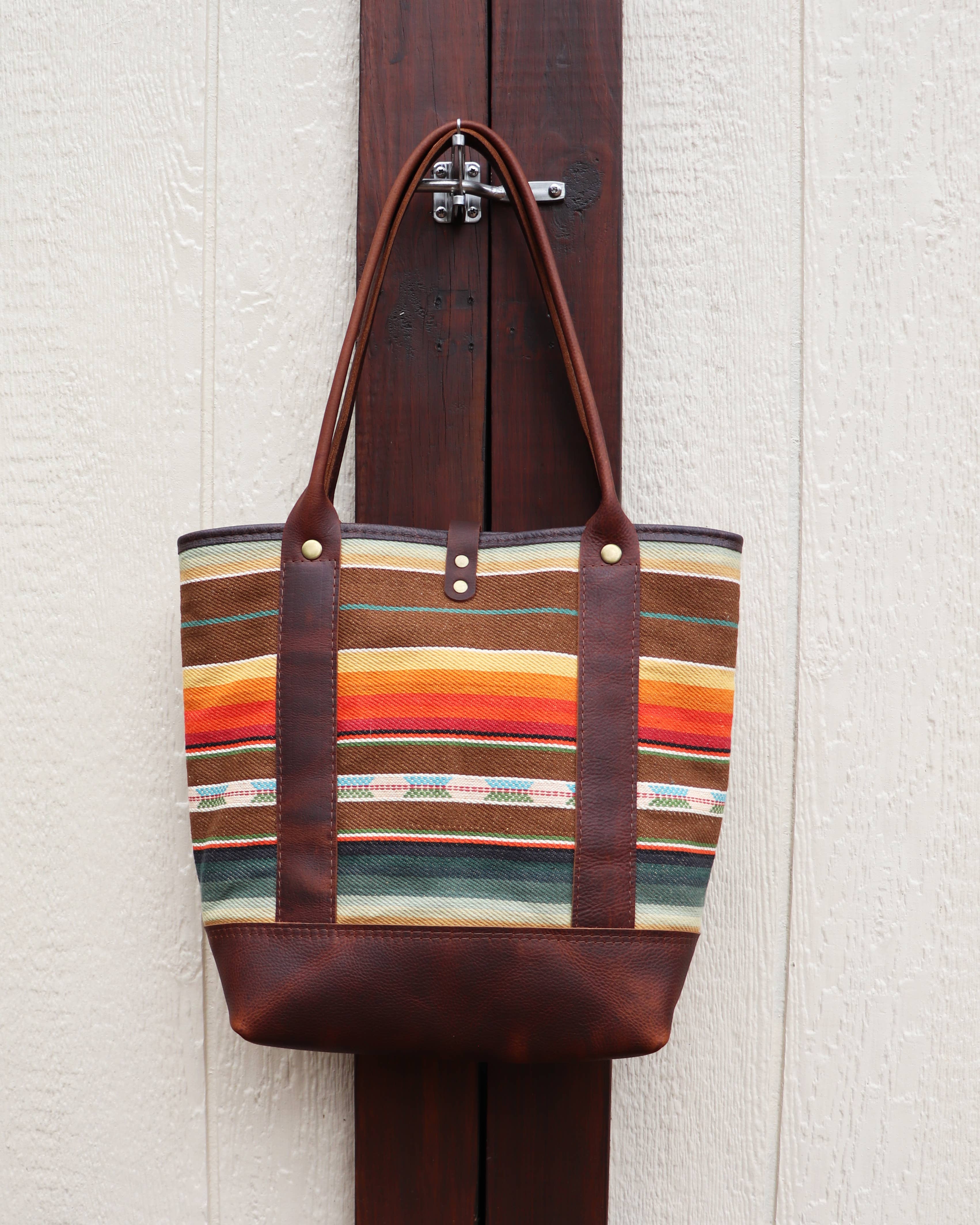 The Pearl Serape Tote by Hawks & Doves, featuring brown full grain leather straps and base with vibrant serape stripes in orange, yellow, green, and beige, is displayed on a wooden door against a light textured wall.