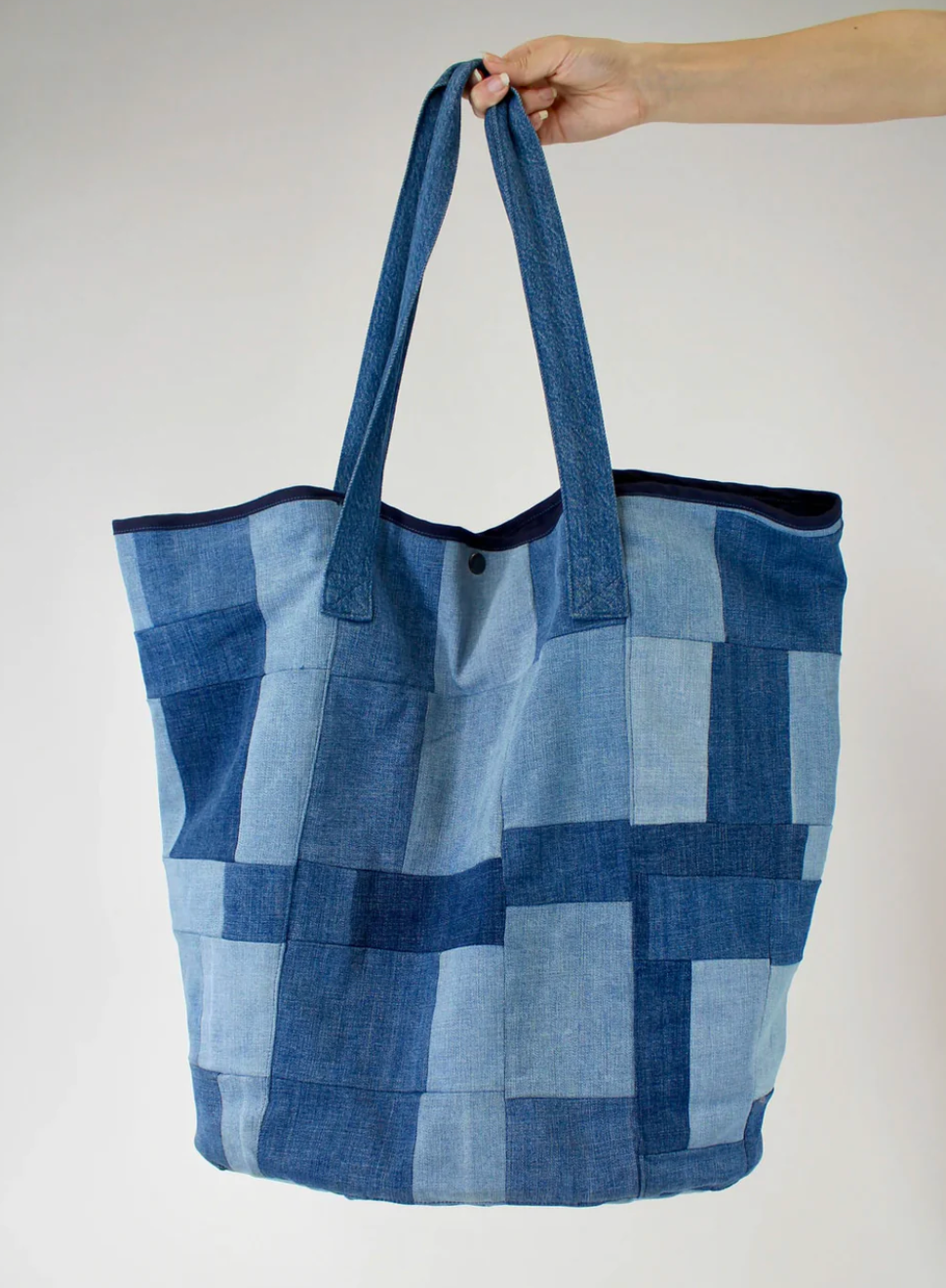 A hand holds the PATCHWORK TOTE by Porter Blue Apparel, a spacious bag crafted from denim squares in blue tones, with long handles and a button closure, set against a plain light background.