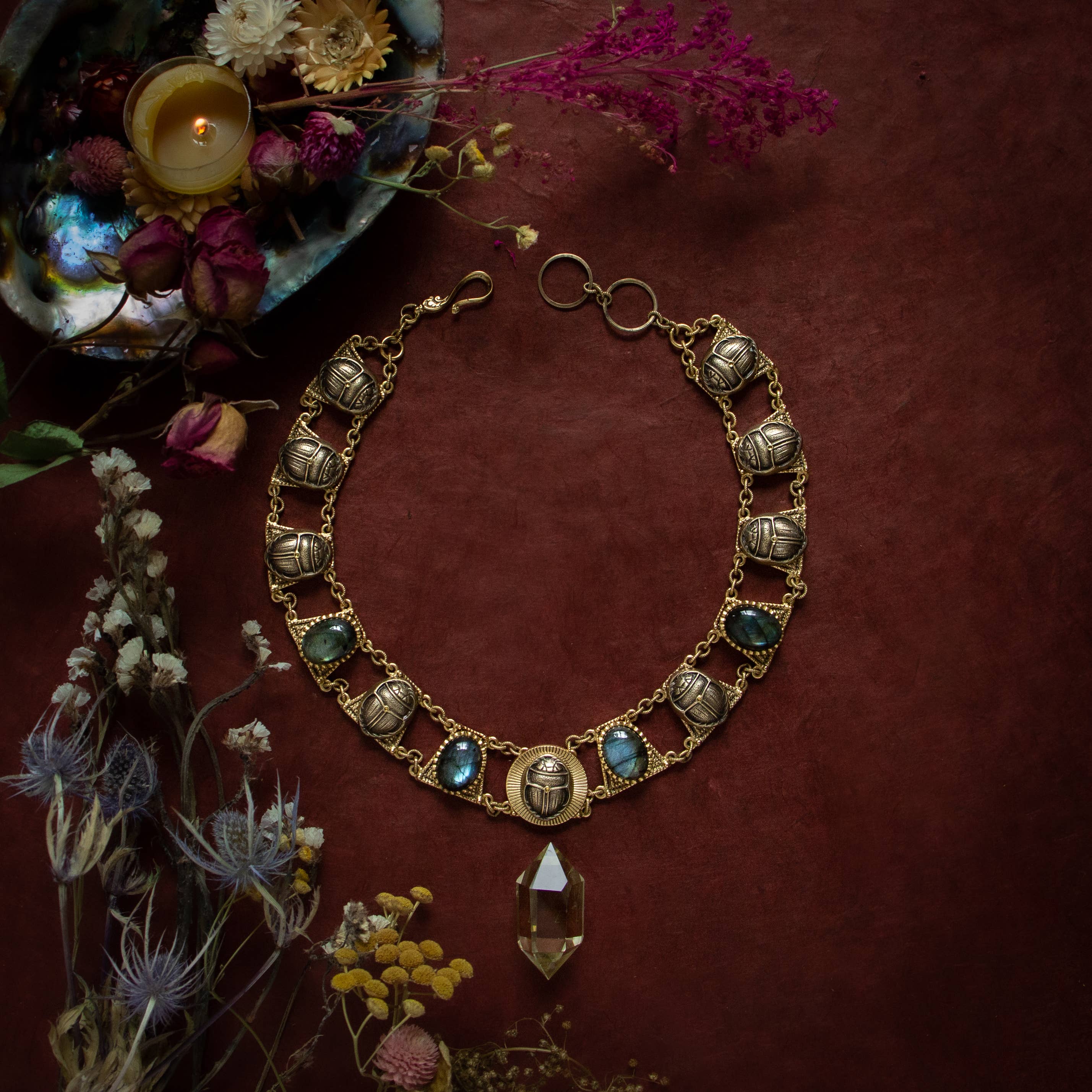 Sacred Scarab Collar Necklace with brass scarab charms and blue stones displayed with dried flowers and a lit candle.