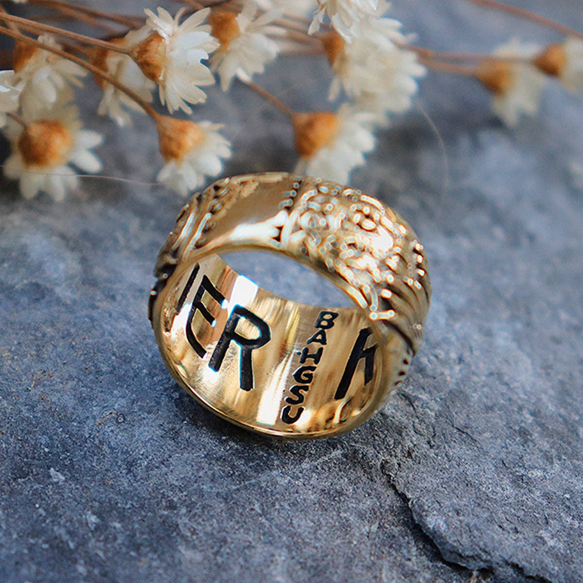 The Rainbow Serpent Ring from bahgsu jewels, a Xavier Rudd collab, rests on a stone surface amid dried white flowers, its gold band featuring intricate designs and the engraved text ER BAHGSU.