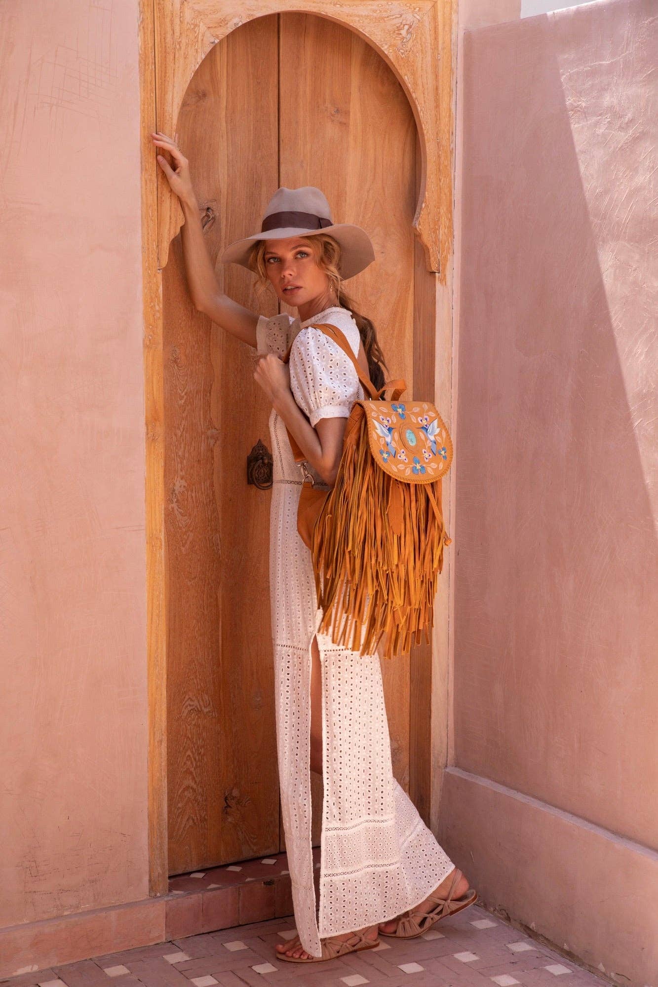 A woman in a white crochet dress, tan hat, and sandals stands by an arched wooden door, carrying the Jodi Lee Monterey Backpack in Antique Tan with blue embroidery, looking over her shoulder at the camera.