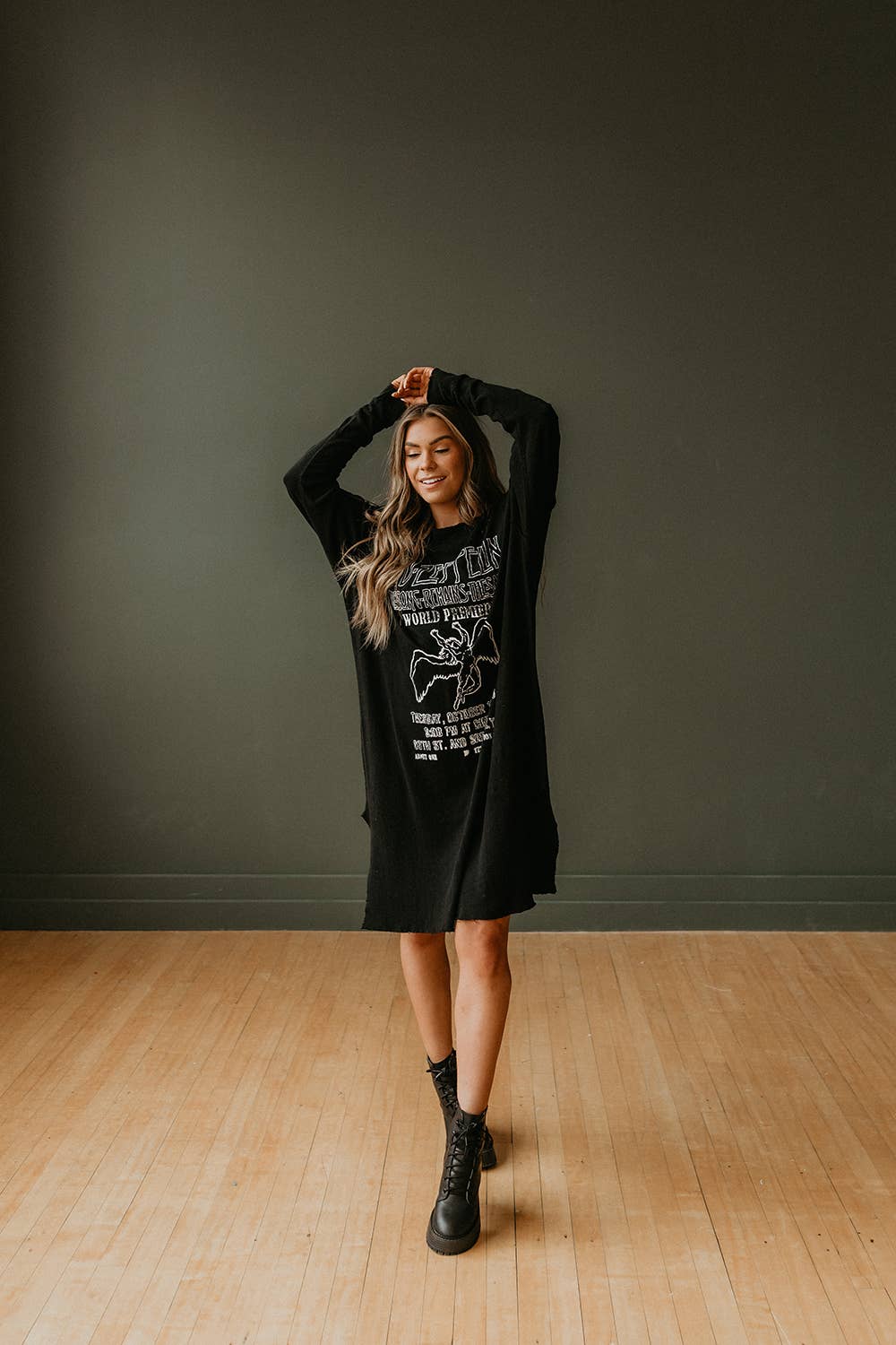 A woman smiles with arms raised, wearing the People Of Leisure Led Zeppelin The Song Remains the Same Dress styled as a long black graphic sweatshirt with black combat boots, standing on a light wooden floor against a dark green wall.