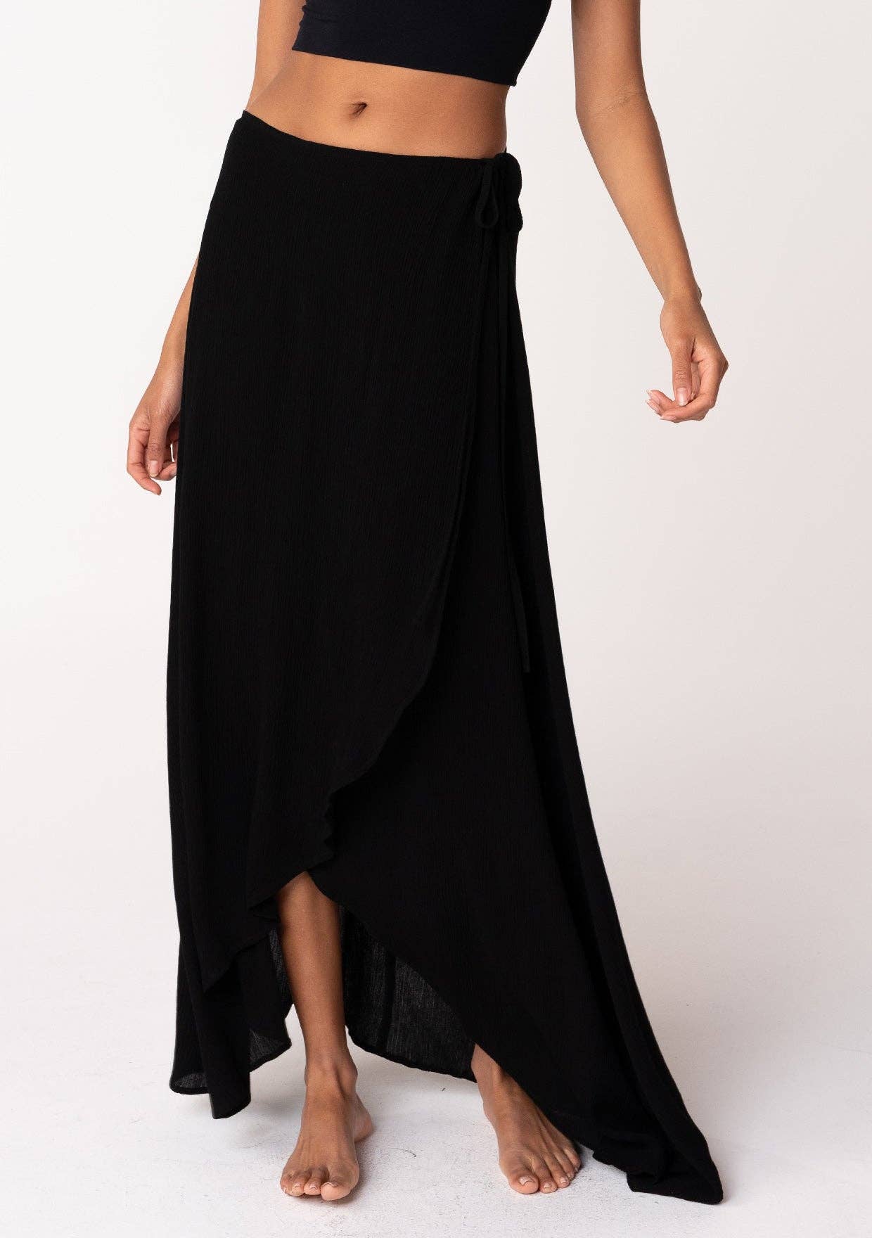 Woman wearing a black Summer Solid Wrap Maxi Skirt with a high-low hem and side tie detail barefoot on white background