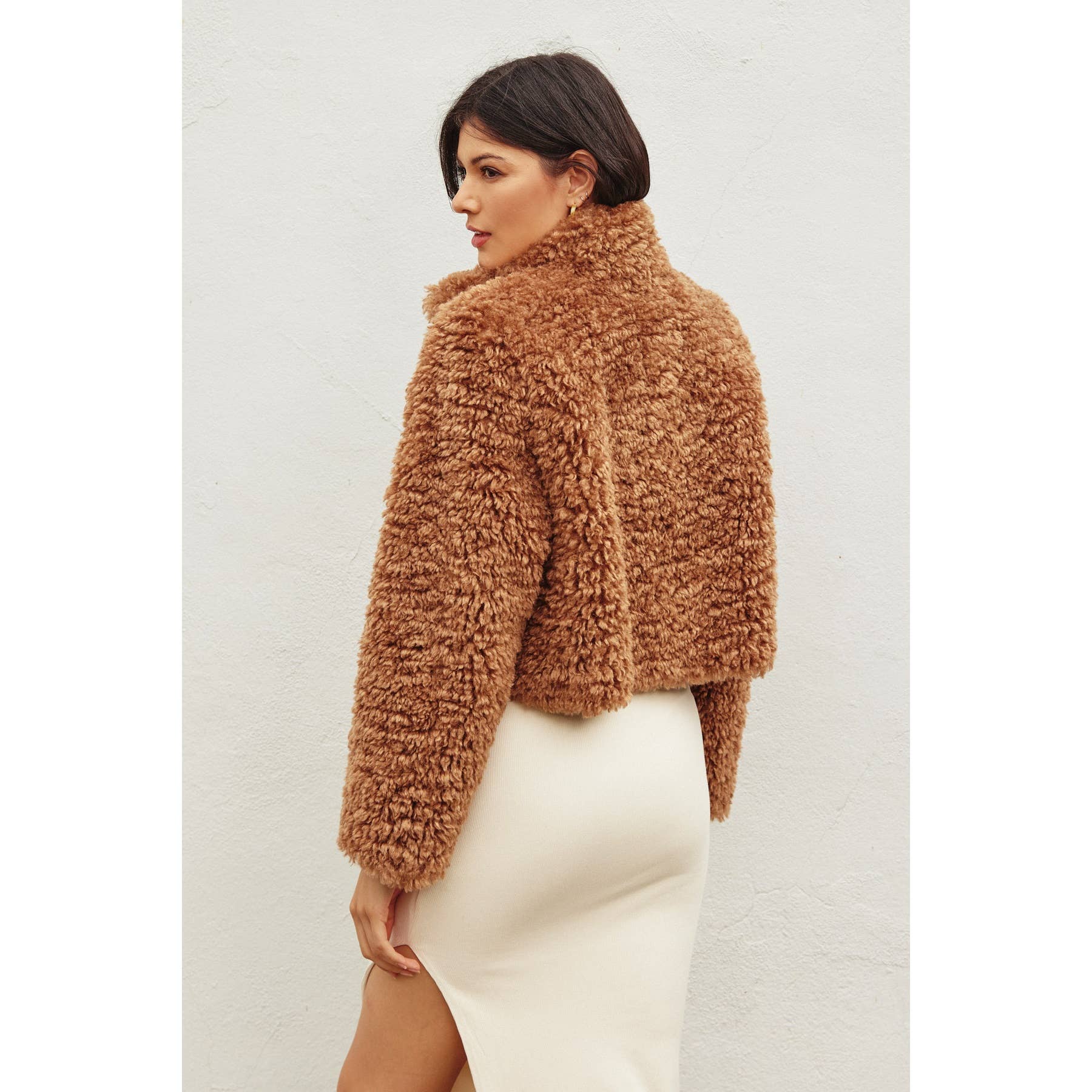 A woman with short dark hair models the Dress Forum Soft Teddy Open Front Cropped Jacket, paired with a white, form-fitting skirt featuring a thigh-high slit, as she stands against a plain light-colored wall.