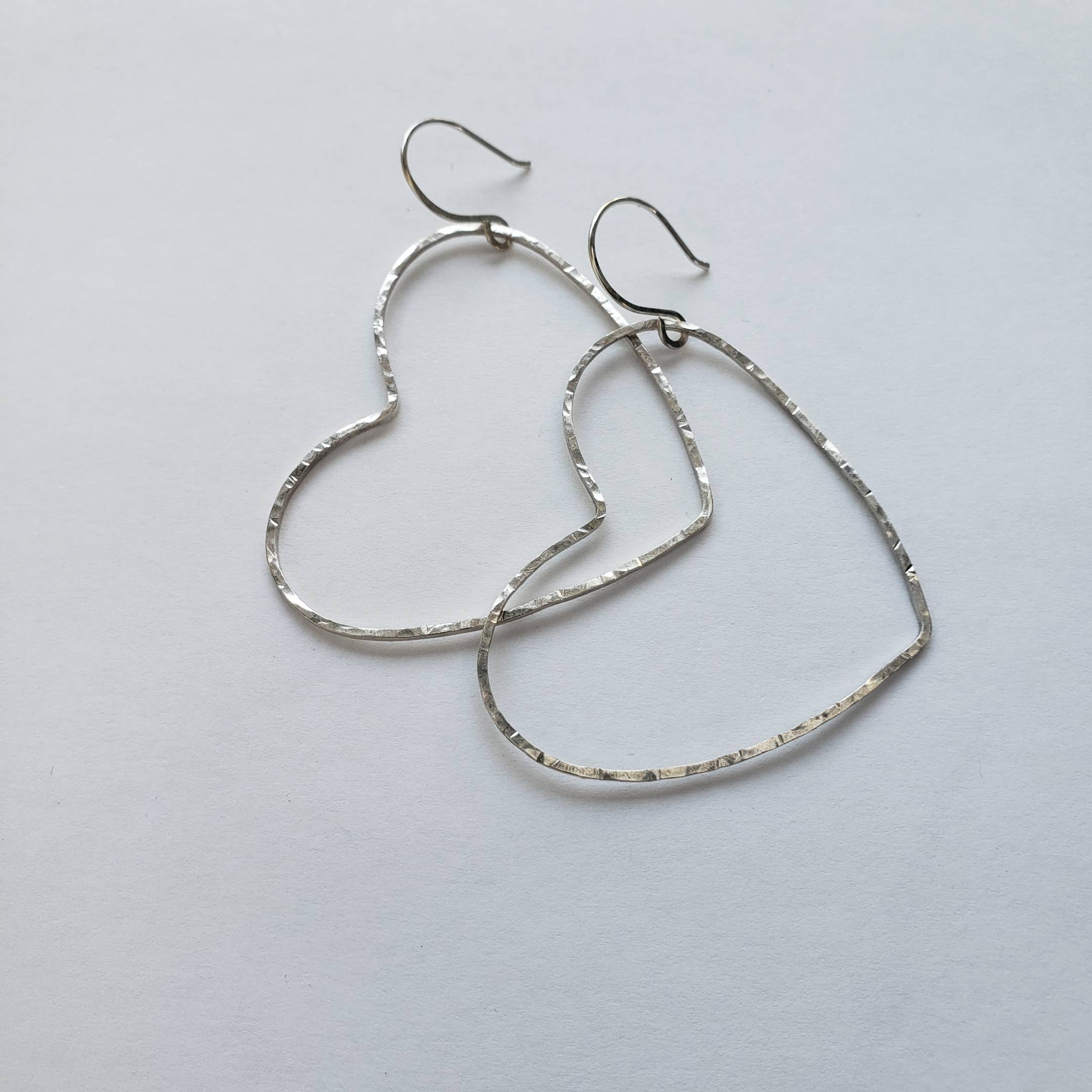 Big Silver Heart Hoop Earrings by Dreamers of Dreams feature hammered silver hearts and sterling ear hooks. Lightweight and handmade, these statement earrings add charm to any outfit on a simple white background.