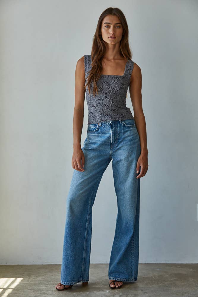 A woman with long brown hair stands confidently against a light wall, wearing the By Together Knit Modal Nylon Spandex Seamless Jacquard Tank, styled with high-waisted wide-leg blue jeans and black sandals.
