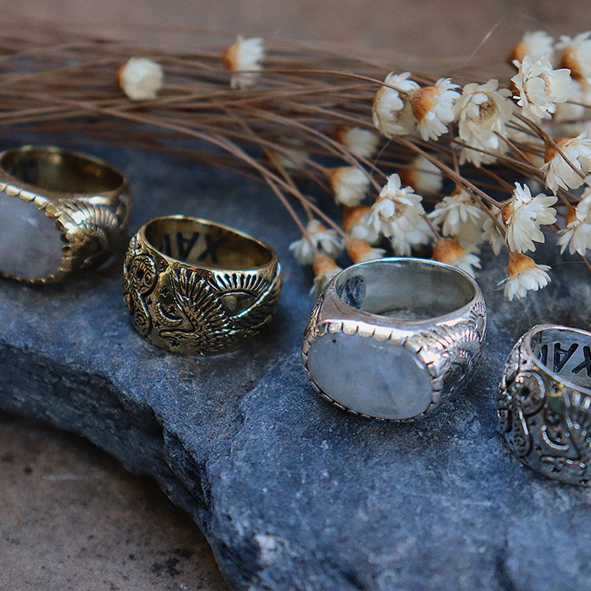 Four ornate rings by bahgsu jewels, including the Rainbow Serpent Ring from the Xavier Rudd Collab, lie on a stone surface with dried white flowers. Each features engraved designs and large oval stones in gold and sterling silver settings.