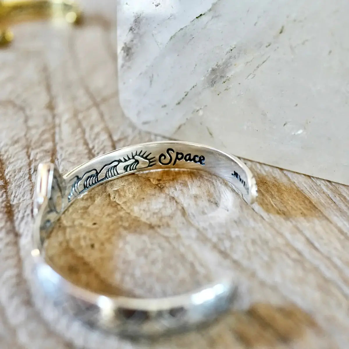 Close-up of a silver Hold Space Cuff bracelet with engraved designs placed on a wooden surface near a white stone.