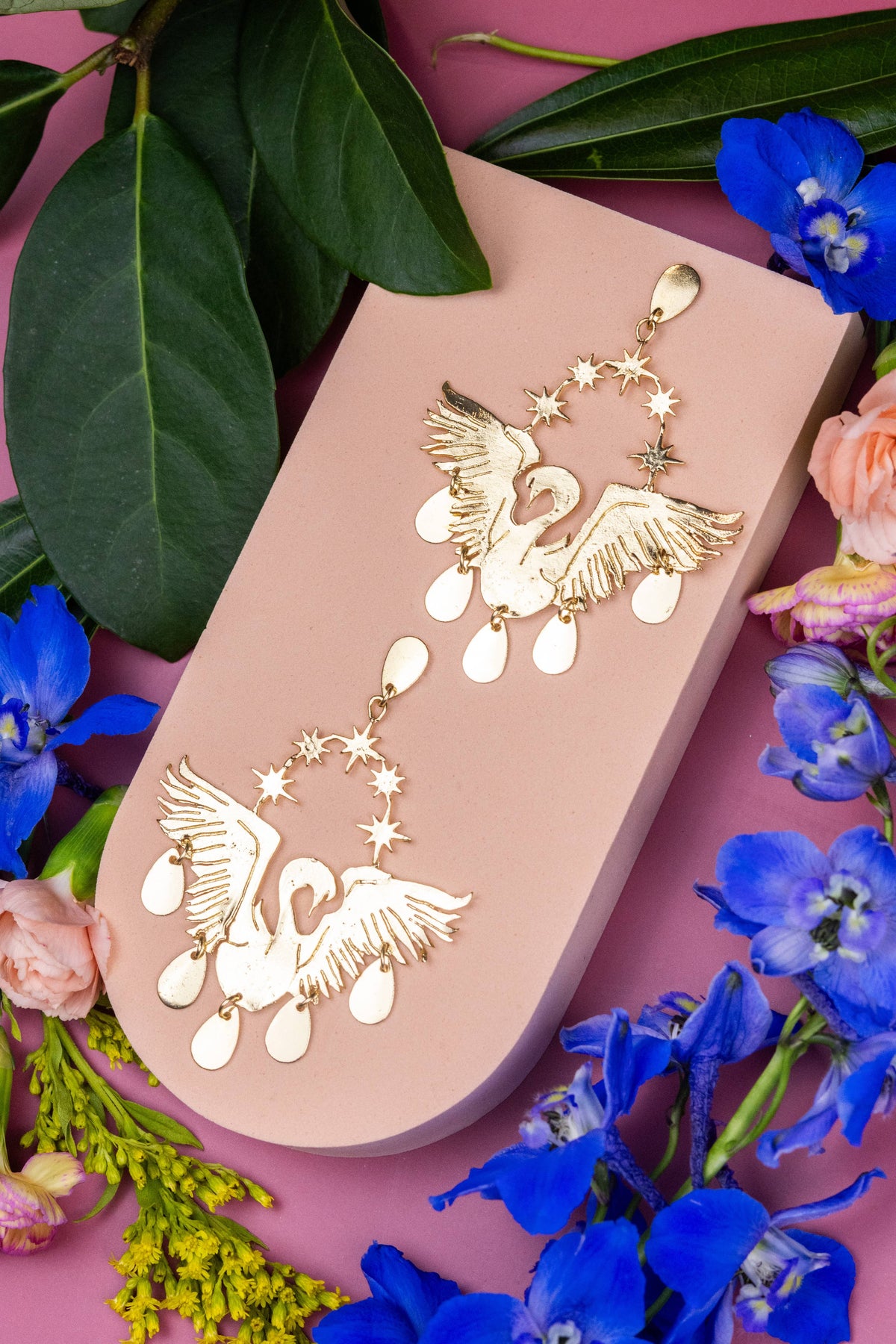 Celestial Swans gold swan earrings on a blush pink display surrounded by colorful flowers