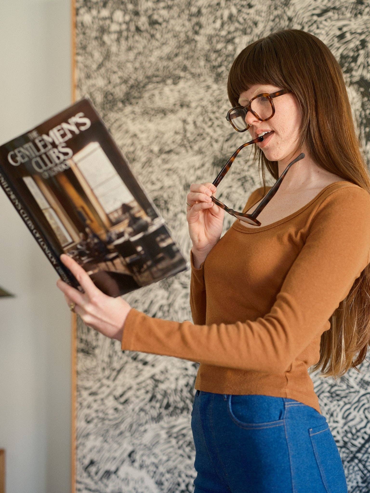 A woman with long brown hair and glasses reads a large book titled Gentlemen’s Clubs while holding another pair of glasses in her mouth. She stands before a patterned wall, wearing blue pants and the Long Sleeve Rib Tee by Mollusk.