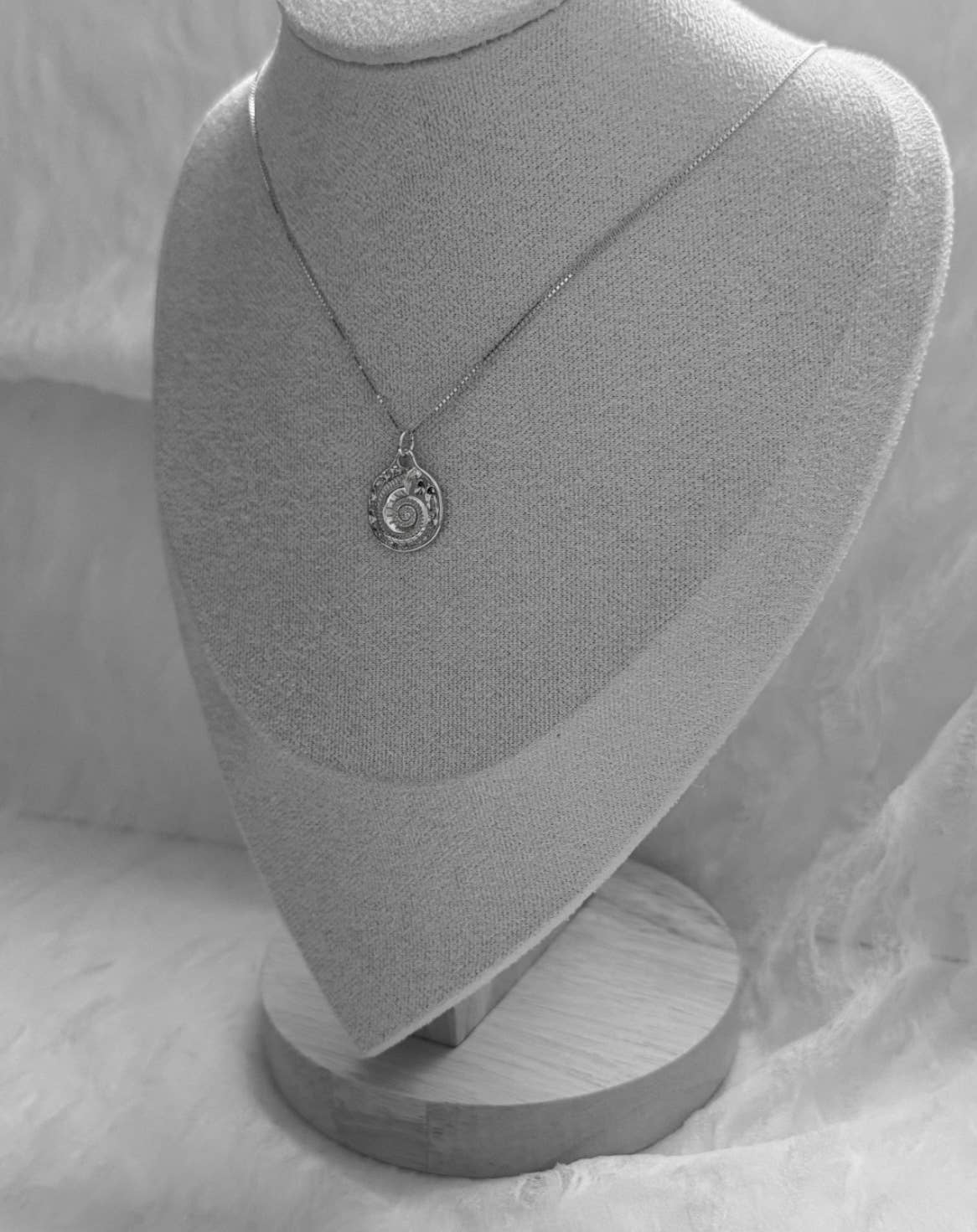 The Mar Medallion Necklace by Tramps+Thieves, a gold vermeil piece with a round jeweled charm, is showcased on a fabric-covered stand against a textured backdrop in this black and white image.