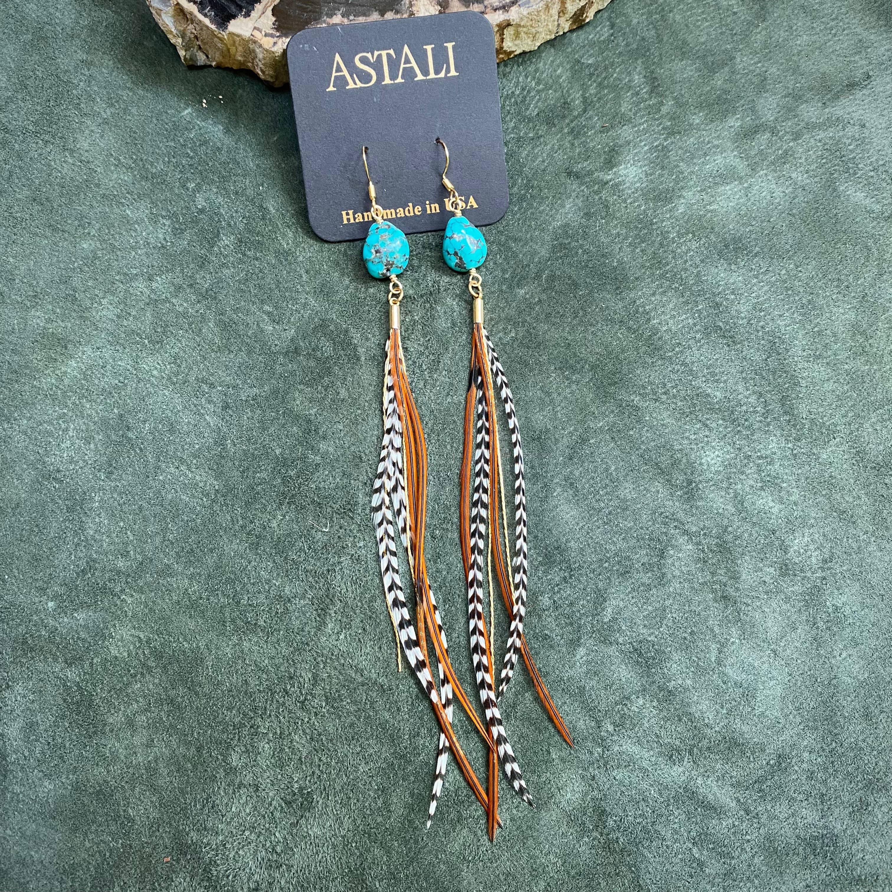 ASTALI’s Turquoise & Mini Feather Earrings – Grizzly & Fawn/Gold, featuring long turquoise stones, brown leather strands, and black-and-white striped feathers, displayed with an ASTALI card on a textured green surface.