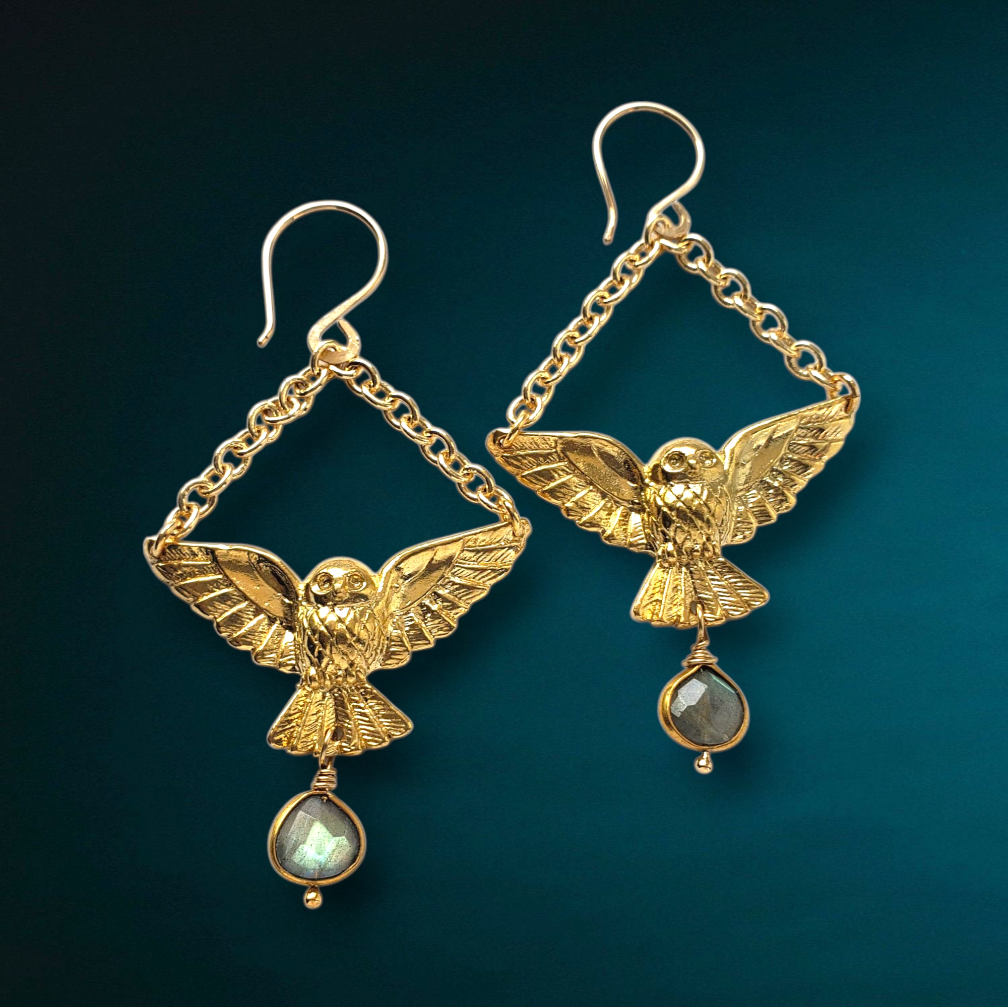 The Gold Owl Labradorite Charm Dangle Earrings by Dreamers of Dreams feature gold-plated owls with outstretched wings, diamond-shaped chains, and round labradorite gems, set against a teal gradient background.