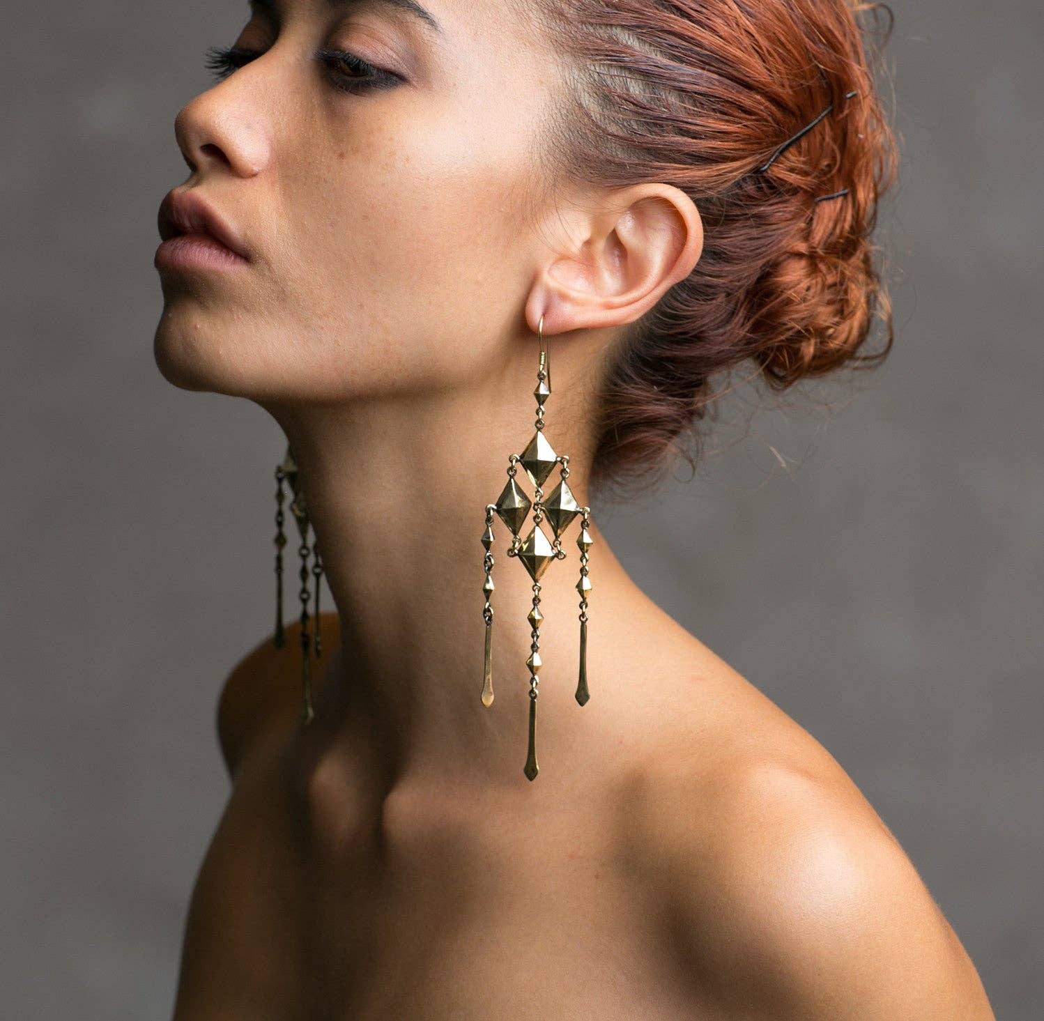 A woman with reddish-brown hair pulled back models the Jan Hilmer + Sparrow Gem chainmail earrings, large and ornate in gold geometric brass, as she turns her head to the side against a plain, neutral background.