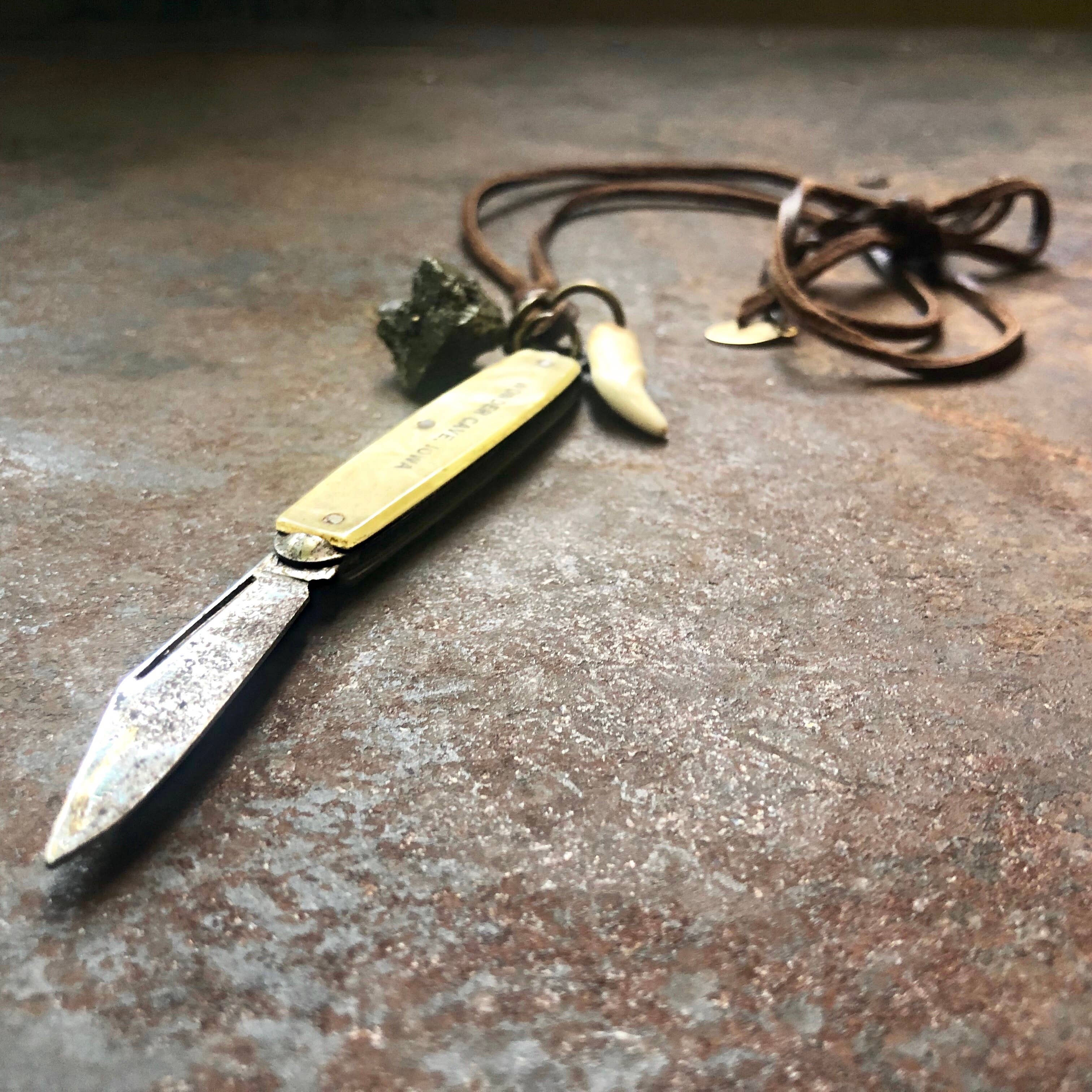 The ASTALI Vintage Pocket Knife & Coyote Tooth Necklace showcases a brass knife, pyrite stone, and coyote tooth on deerskin leather, displayed on a rustic surface.