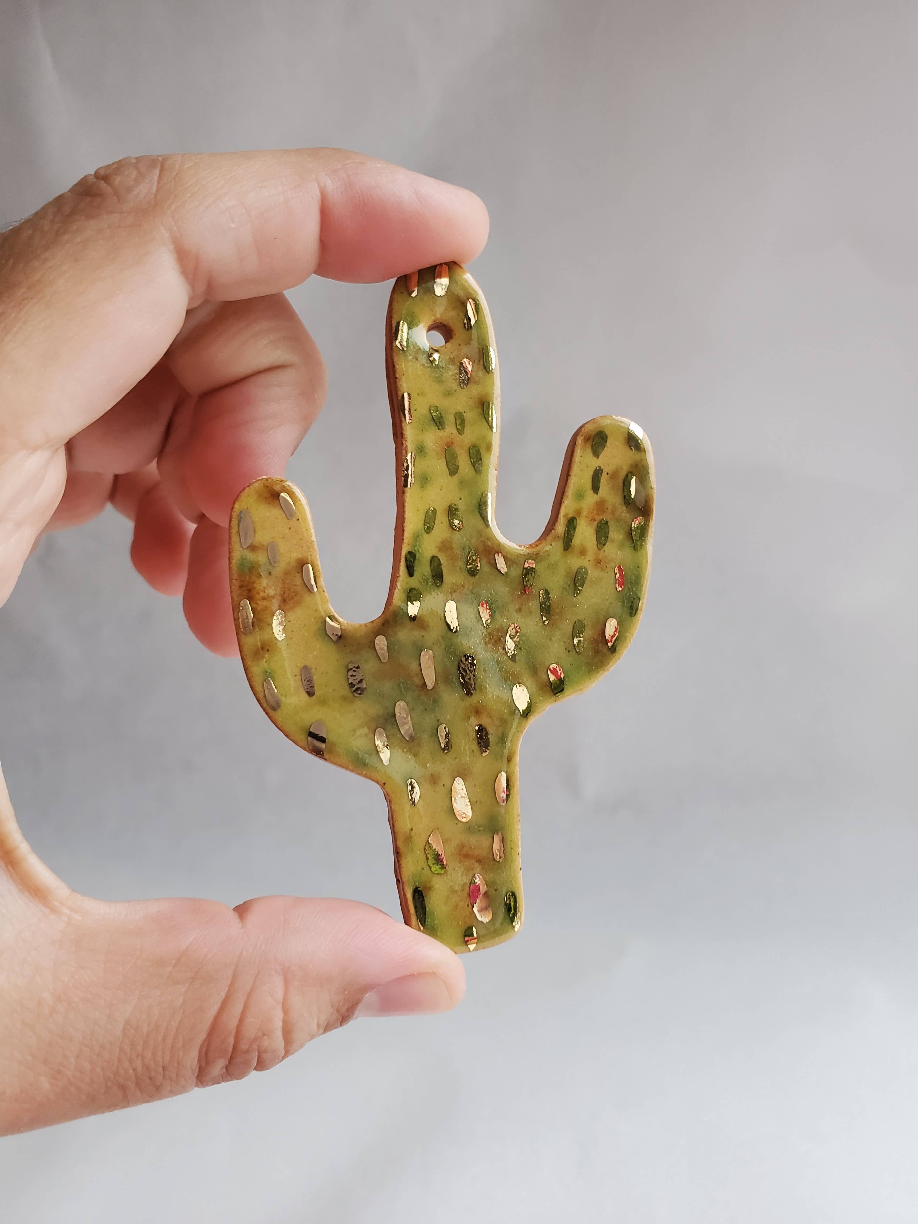Hand holding a small ceramic green cactus ornament with gold accents on a neutral background