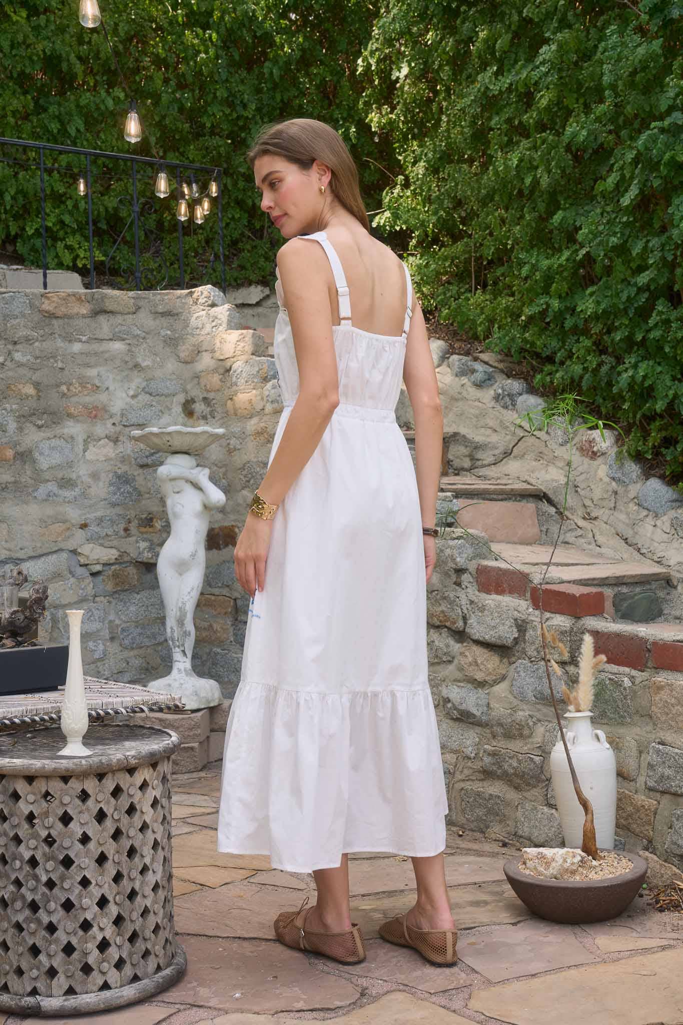 Woman wearing The Molly Embroidered Detail Midi Dress in white, standing outdoors with greenery and stone walls behind her
