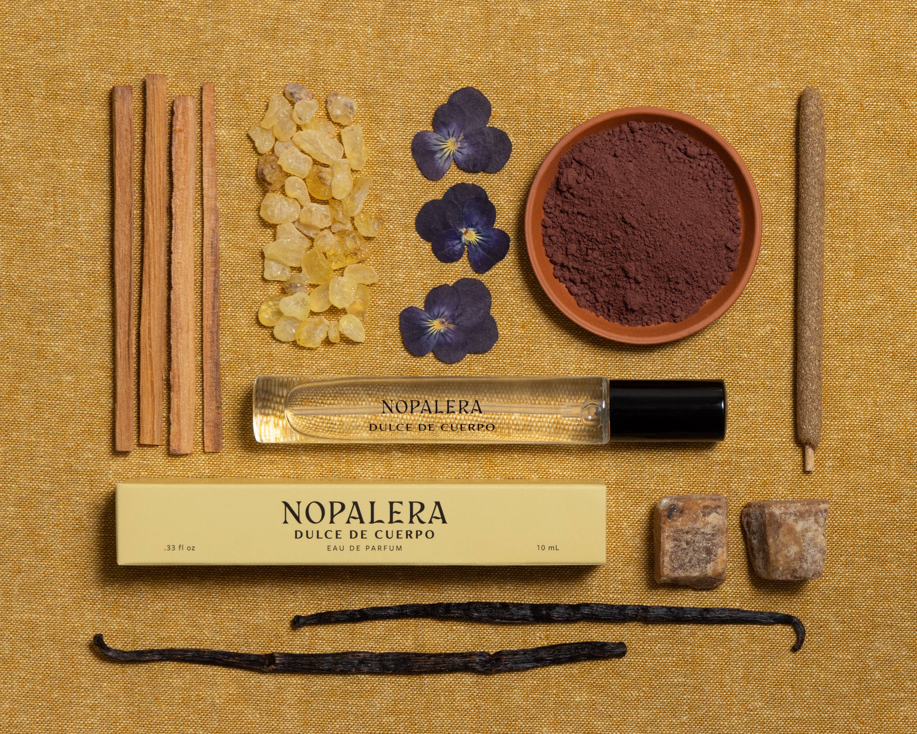 A flat lay of Nopalera’s Dulce de Cuerpo Eau de Parfum – 10mL Travel Spray with its box, surrounded by cinnamon sticks, resin crystals, purple flowers, brown powder, cubes, and vanilla beans evokes a sandalwood-vanilla scent on gold fabric.